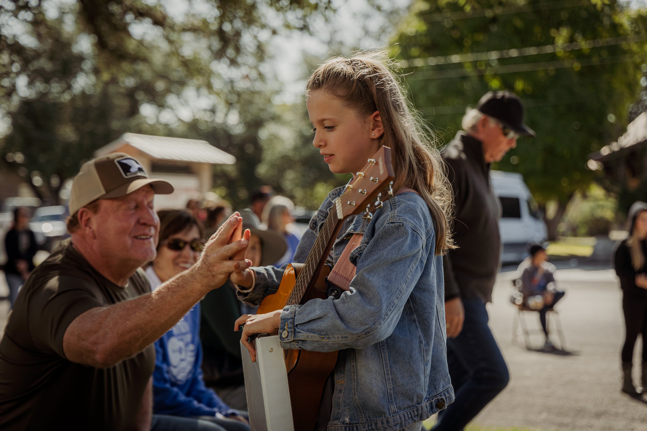 A young girl is receiving a high five from an older man during an outdoor event, with a guitar on her back and people sitting and standing around in a park-like setting.