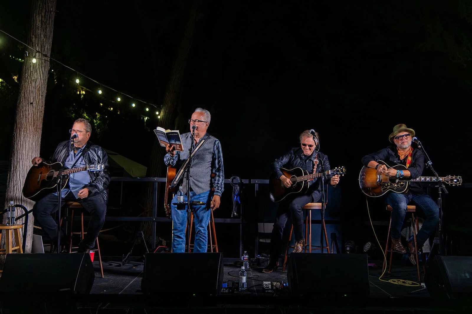 Four musicians performing on stage at night with string lights overhead. The band includes two guitarists, a vocalist, and a bass player, all seated or standing with their instruments.