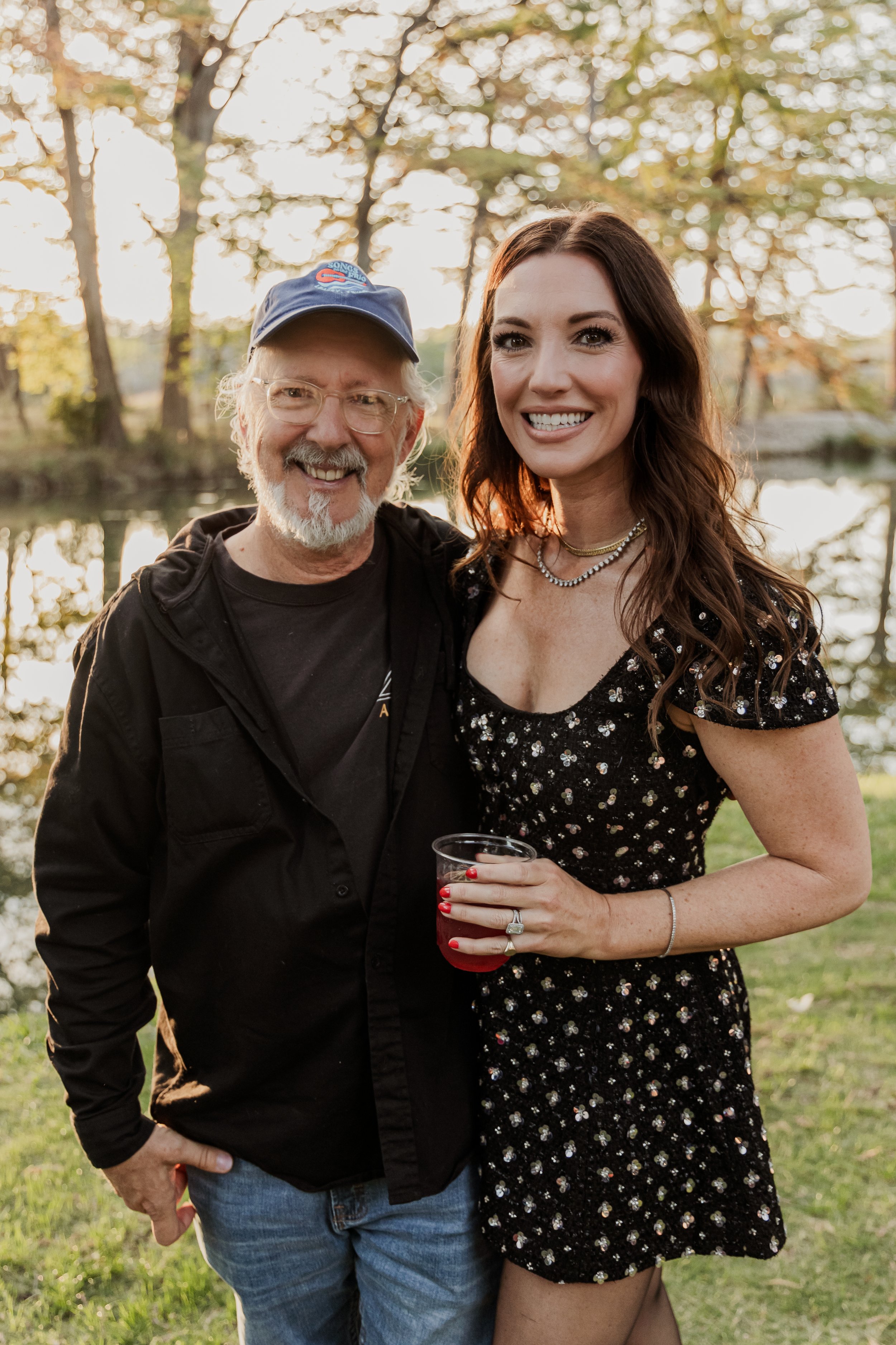 A man and woman smiling outdoors near a body of water with trees in the background, the woman holding a drink.