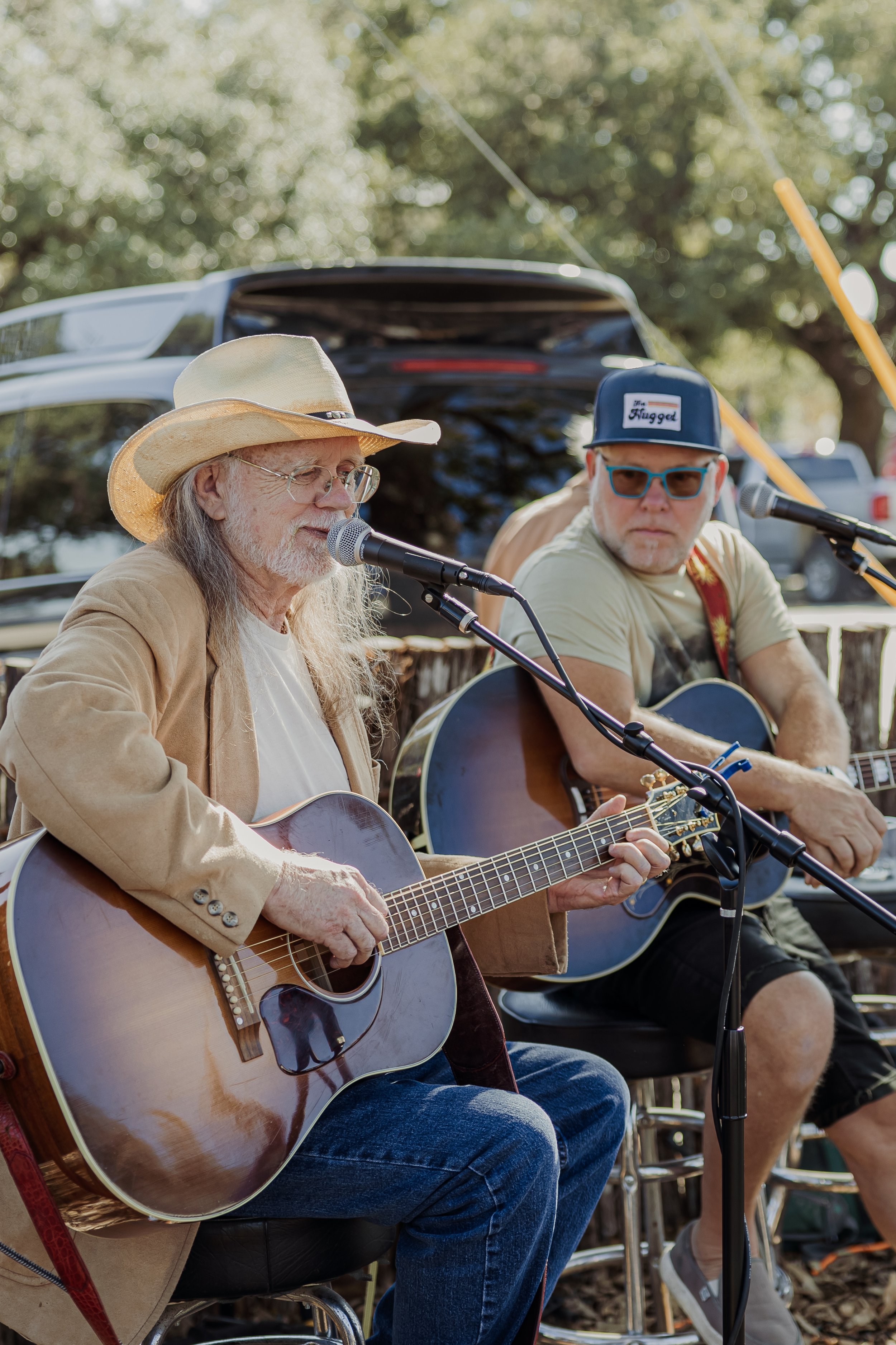 Two men playing guitars and singing outdoors with trees and vehicles in the background.