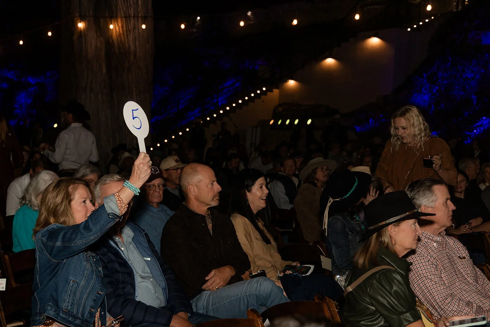 Audience seated in a dark venue, with a woman raising a paddle numbered 5, and others smiling and facing forward.