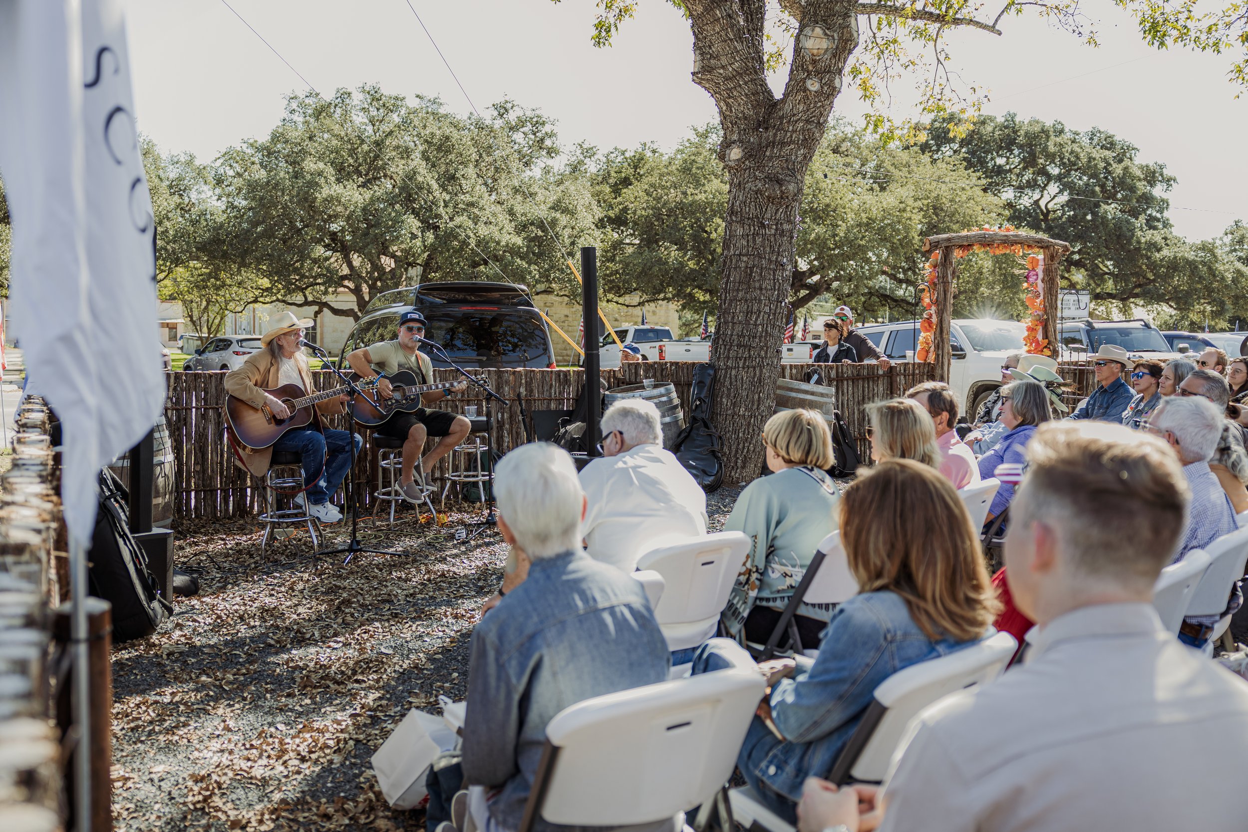 An outdoor gathering with a musical performance, featuring two musicians playing guitars and singing to an audience seated in white chairs under trees.