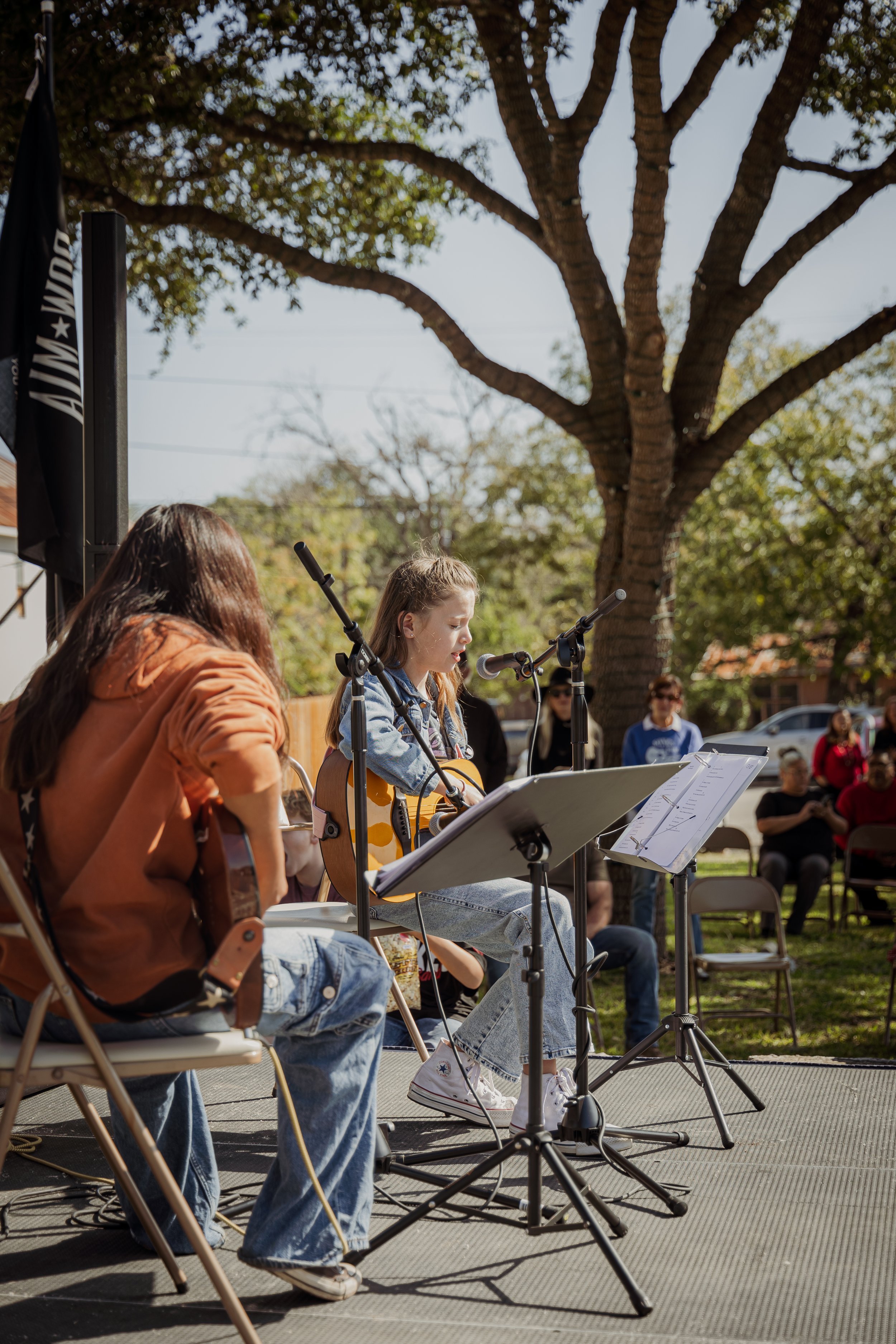 A girl playing acoustic guitar and singing into a microphone during an outdoor music performance, with people sitting and standing in the background under a large tree.