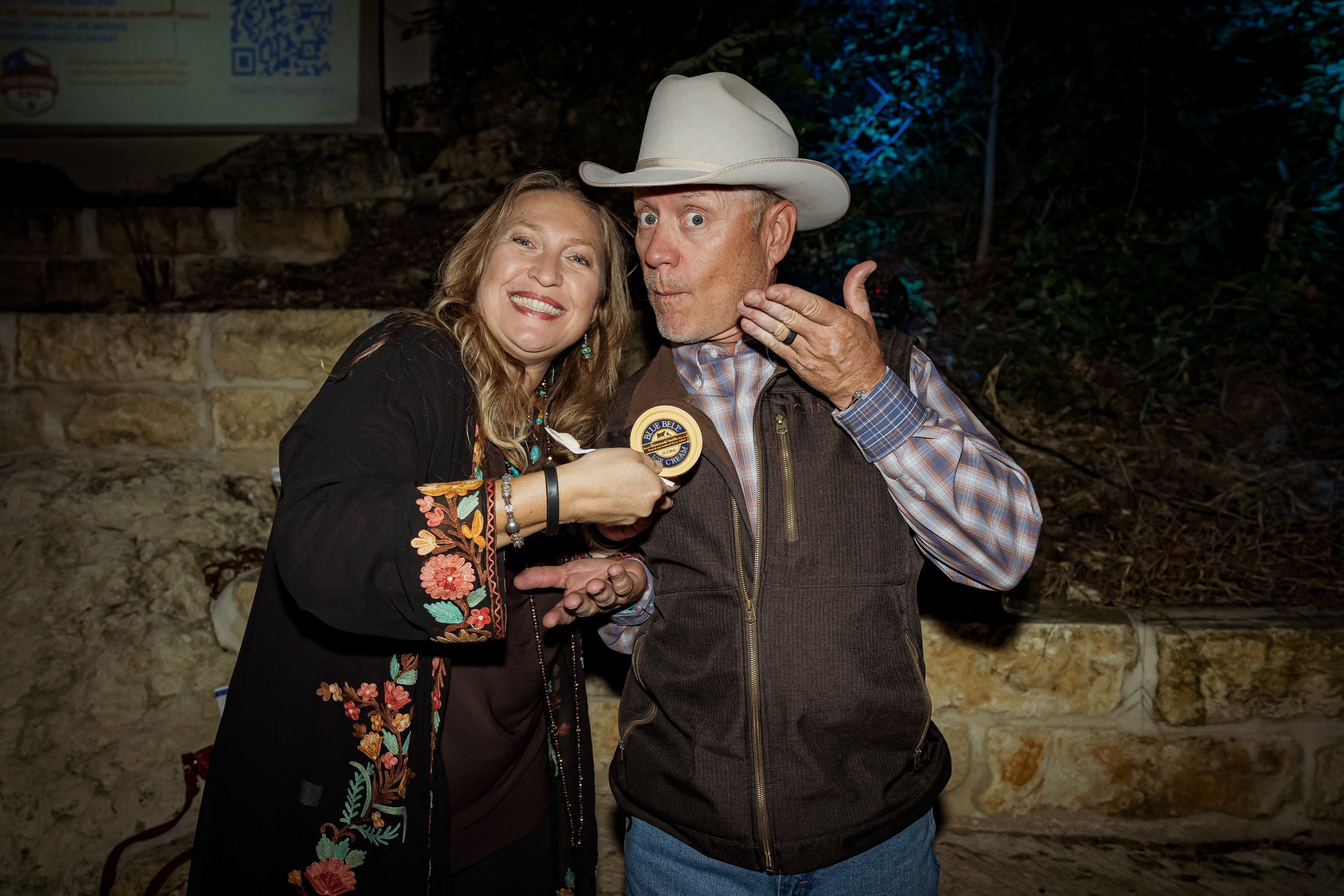 A woman giving a blue ribbon to a man in a cowboy hat, both smiling, outdoors at night with a stone wall and trees in the background.