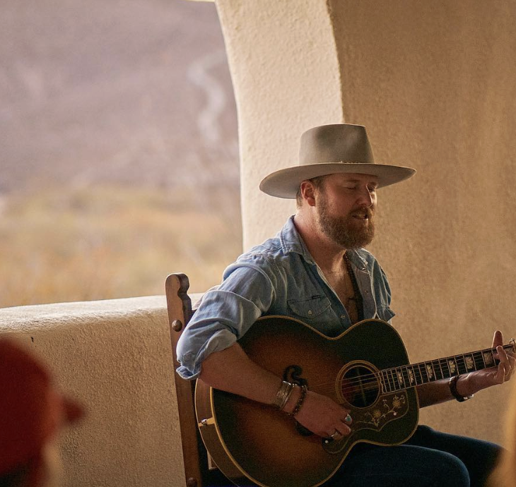 A man with a beard wearing a wide-brimmed hat and a denim shirt playing an acoustic guitar in a cozy indoor setting.