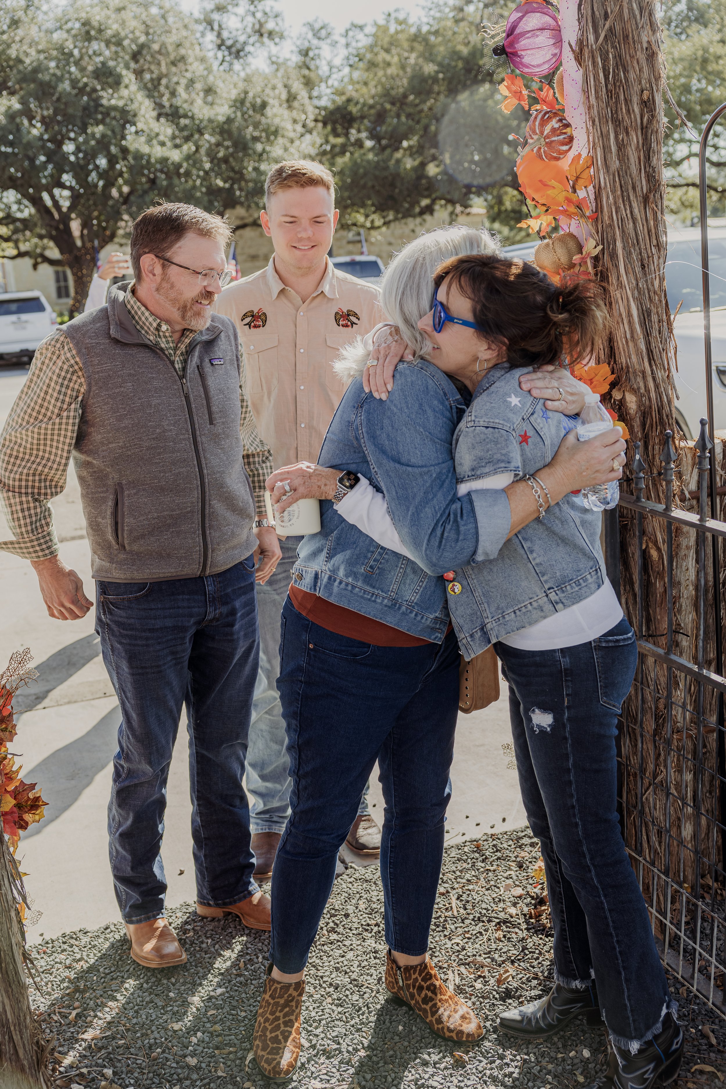 Four people are gathered outdoors, sharing hugs and smiling, near a decorated wooden post with autumn embellishments.