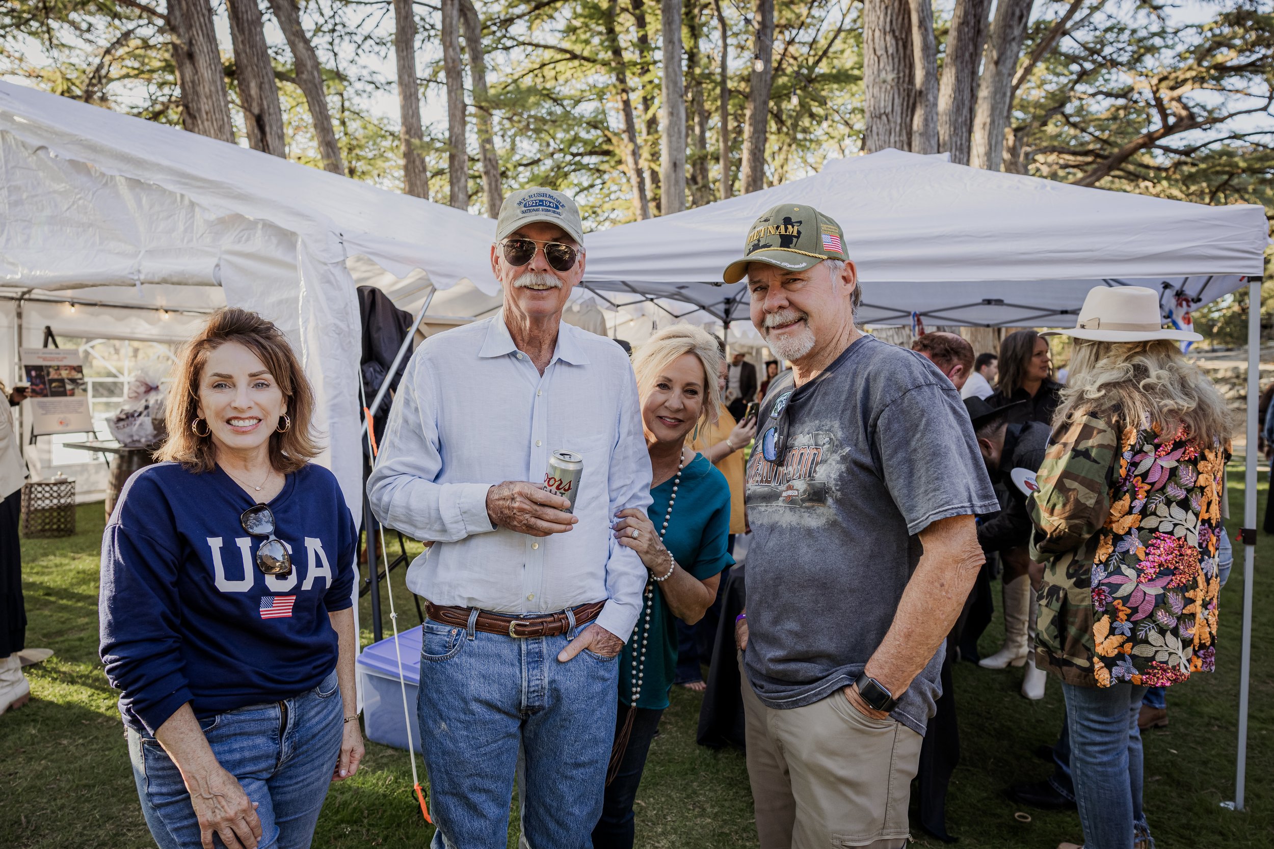Group of five people standing outdoors at a social gathering, smiling, with tents and other people in the background.