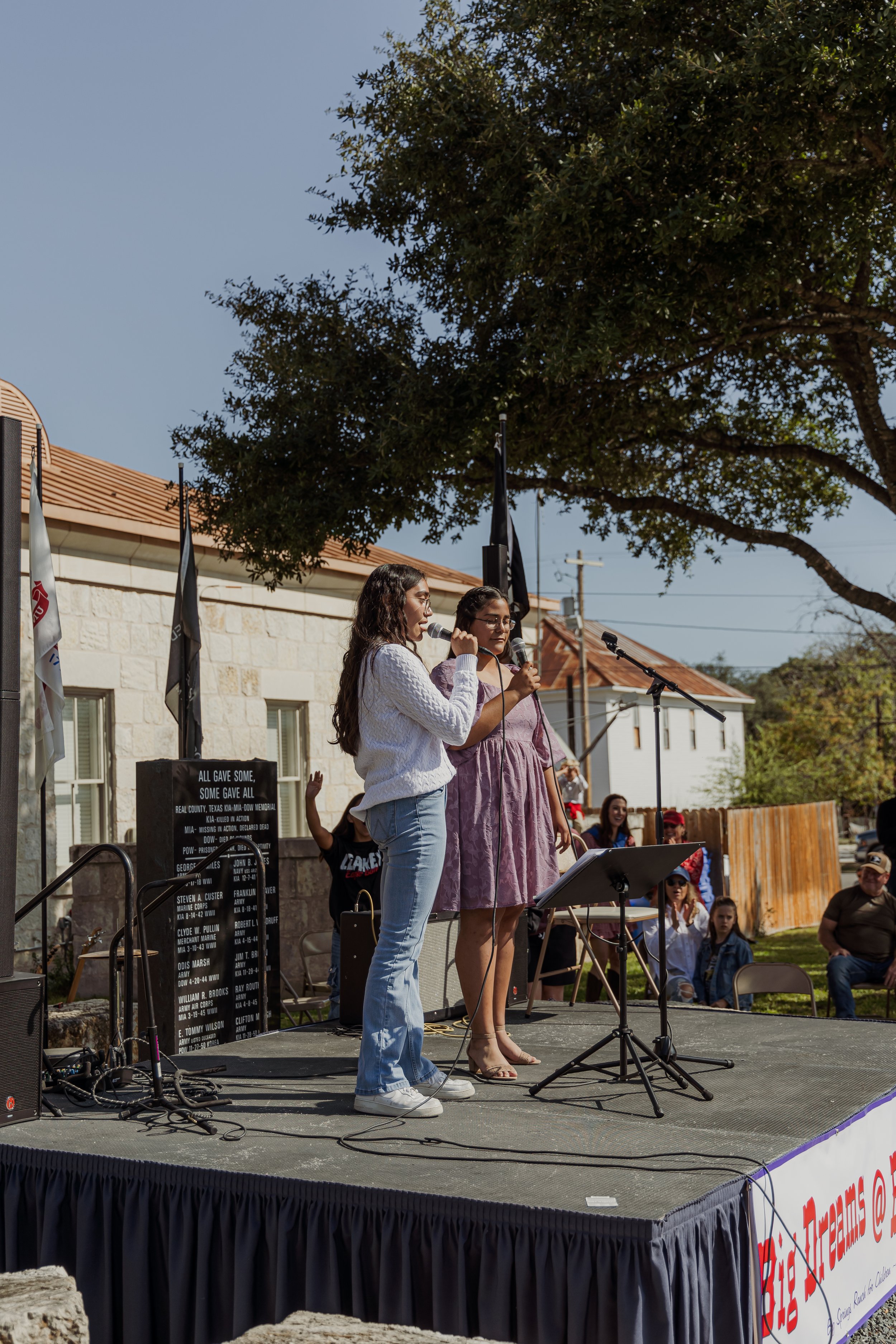 Two women are singing into microphones on an outdoor stage during a community event, with several people and flags in the background.