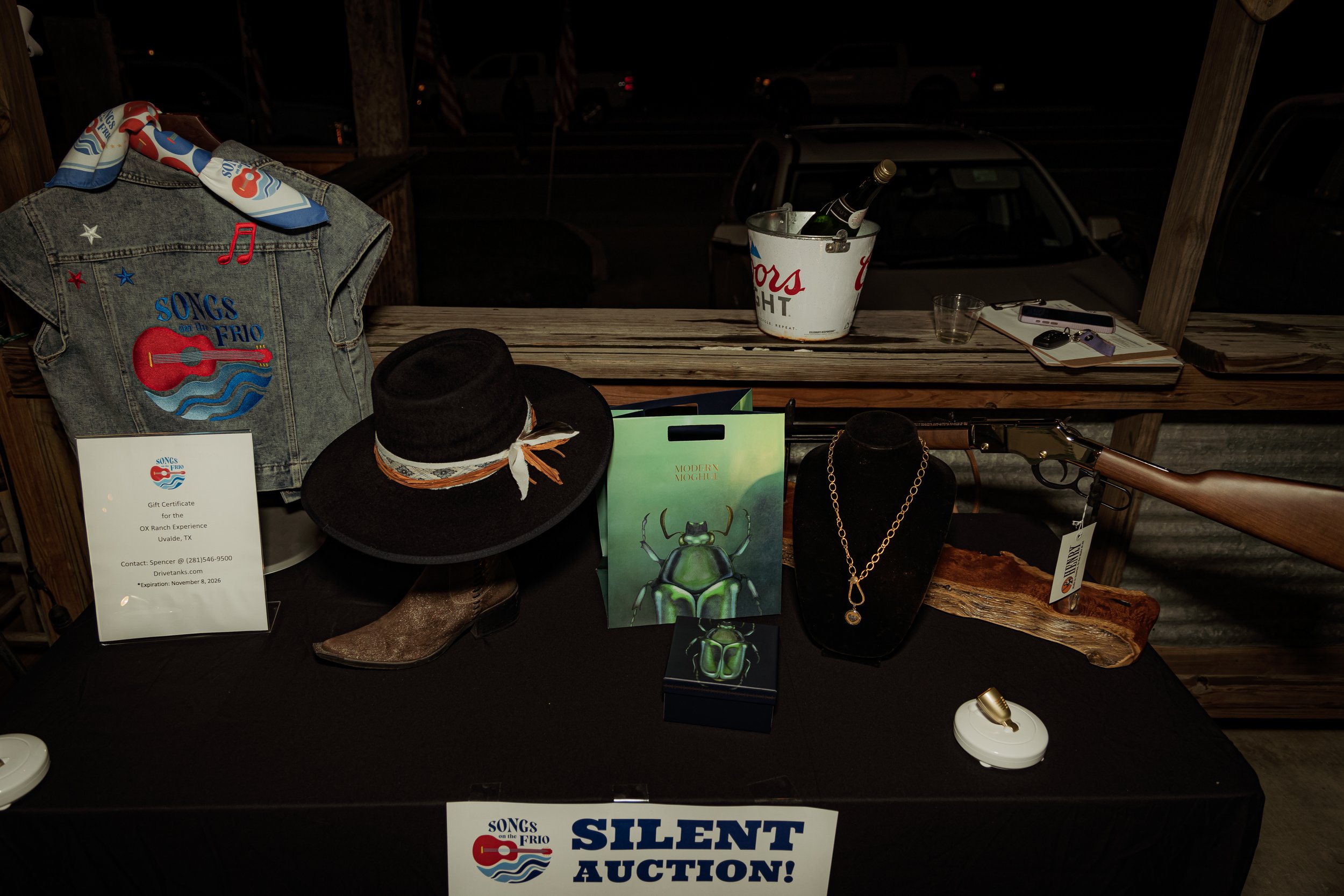 Table display of jewelry, a cowboy hat, a denim vest with 'Songs on the Frio' embroidery, a toy gun, and auction signs, set against a wooden background at night.