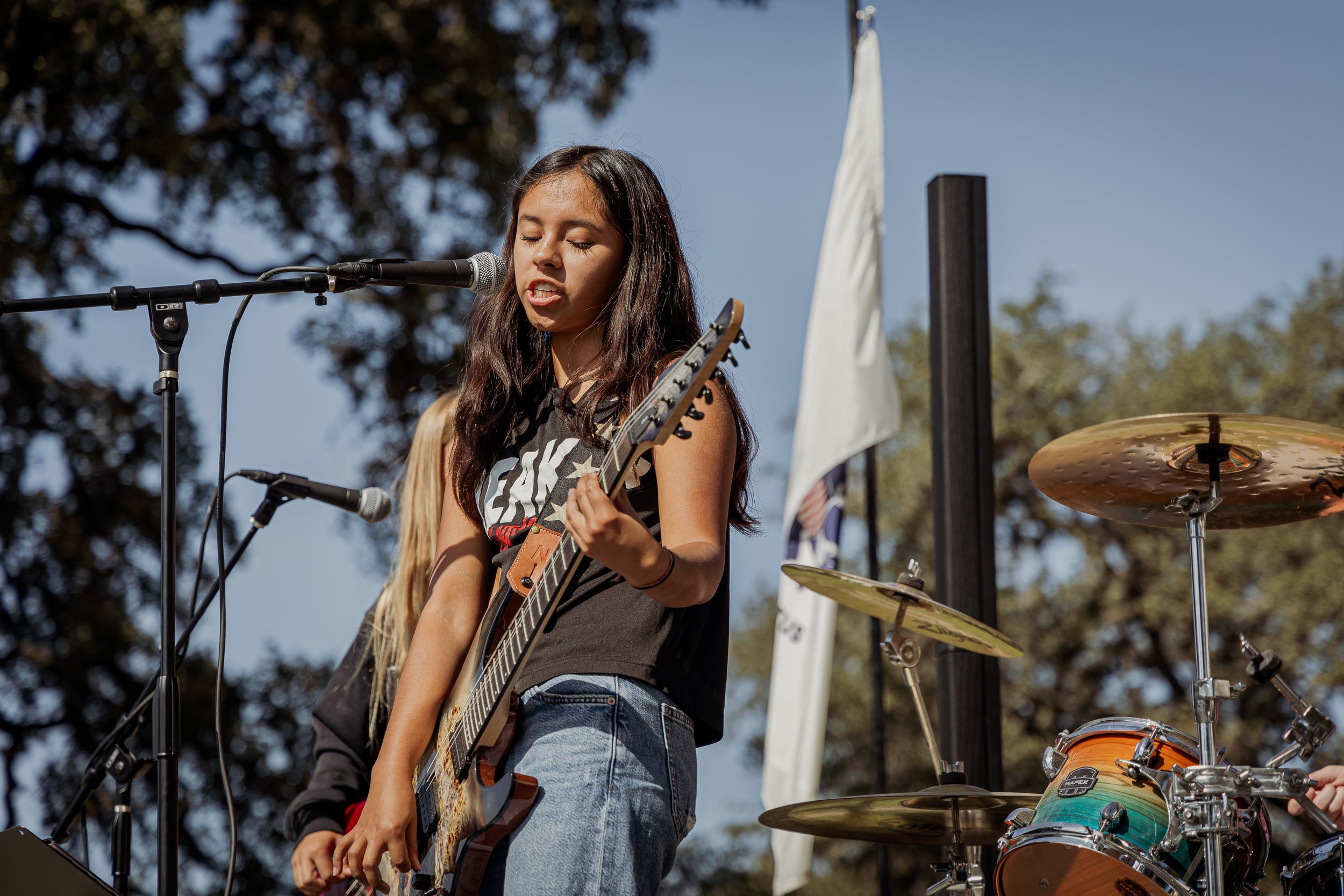 A girl with long dark hair singing into a microphone while playing an electric guitar during an outdoor performance with drumming equipment visible in the background.