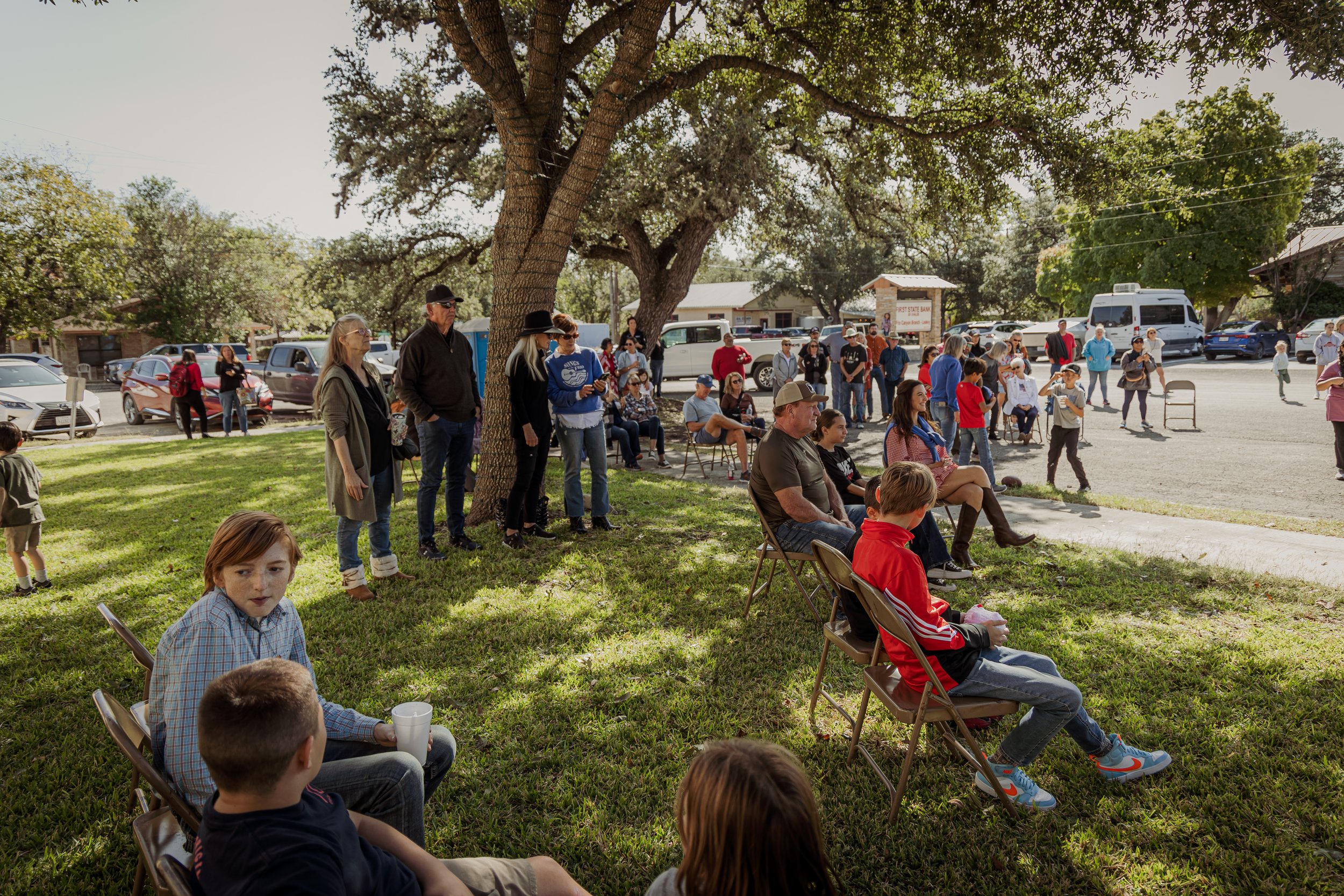 A group of people gathered outdoors in a park-like setting, some sitting on folding chairs under a large tree, others standing around, with cars parked in the background.