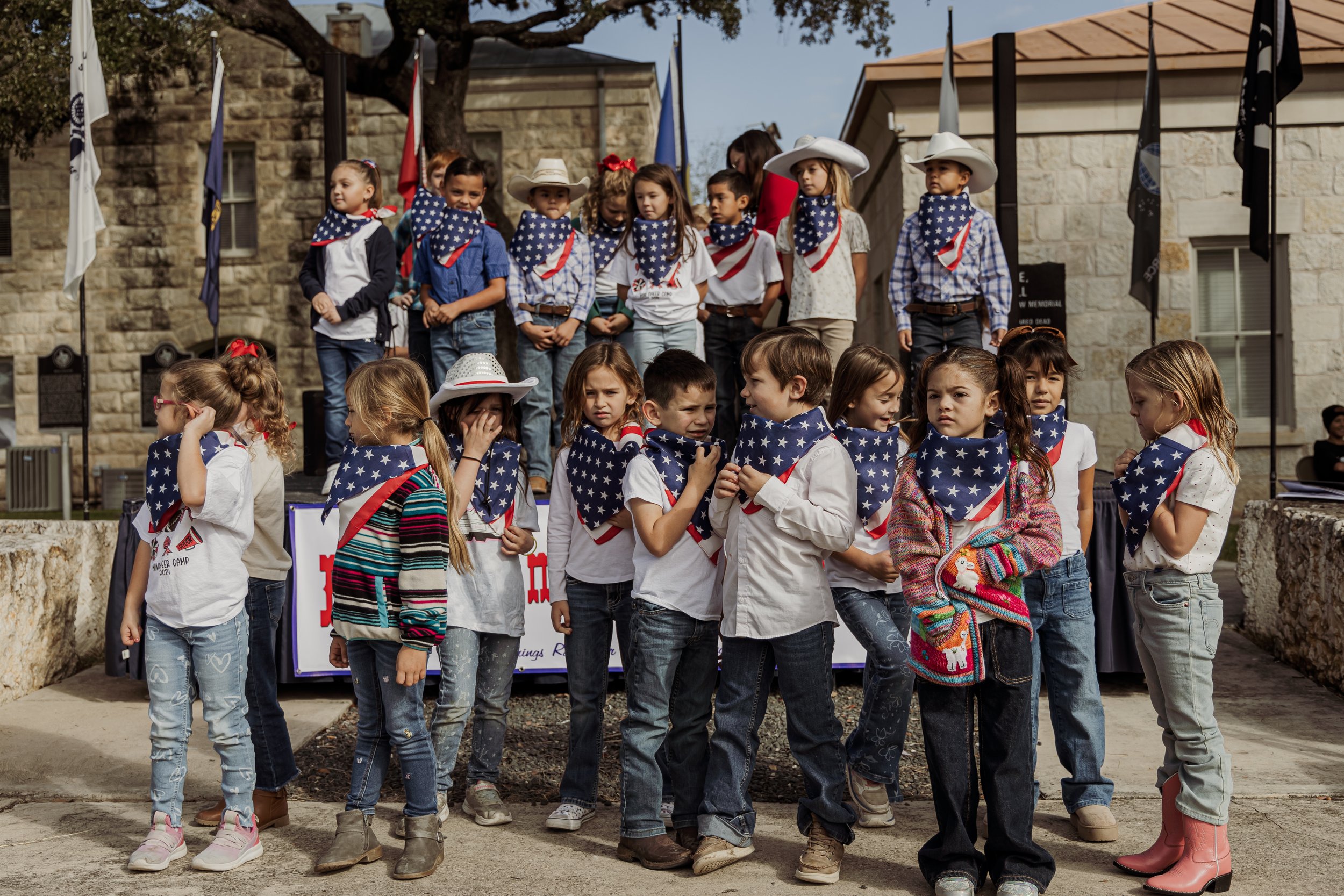 Group of children dressed in patriotic American-themed clothing, some wearing cowboy hats and bandanas with stars and stripes, gathered outdoors during a celebration, with some standing on a stage and others in front.