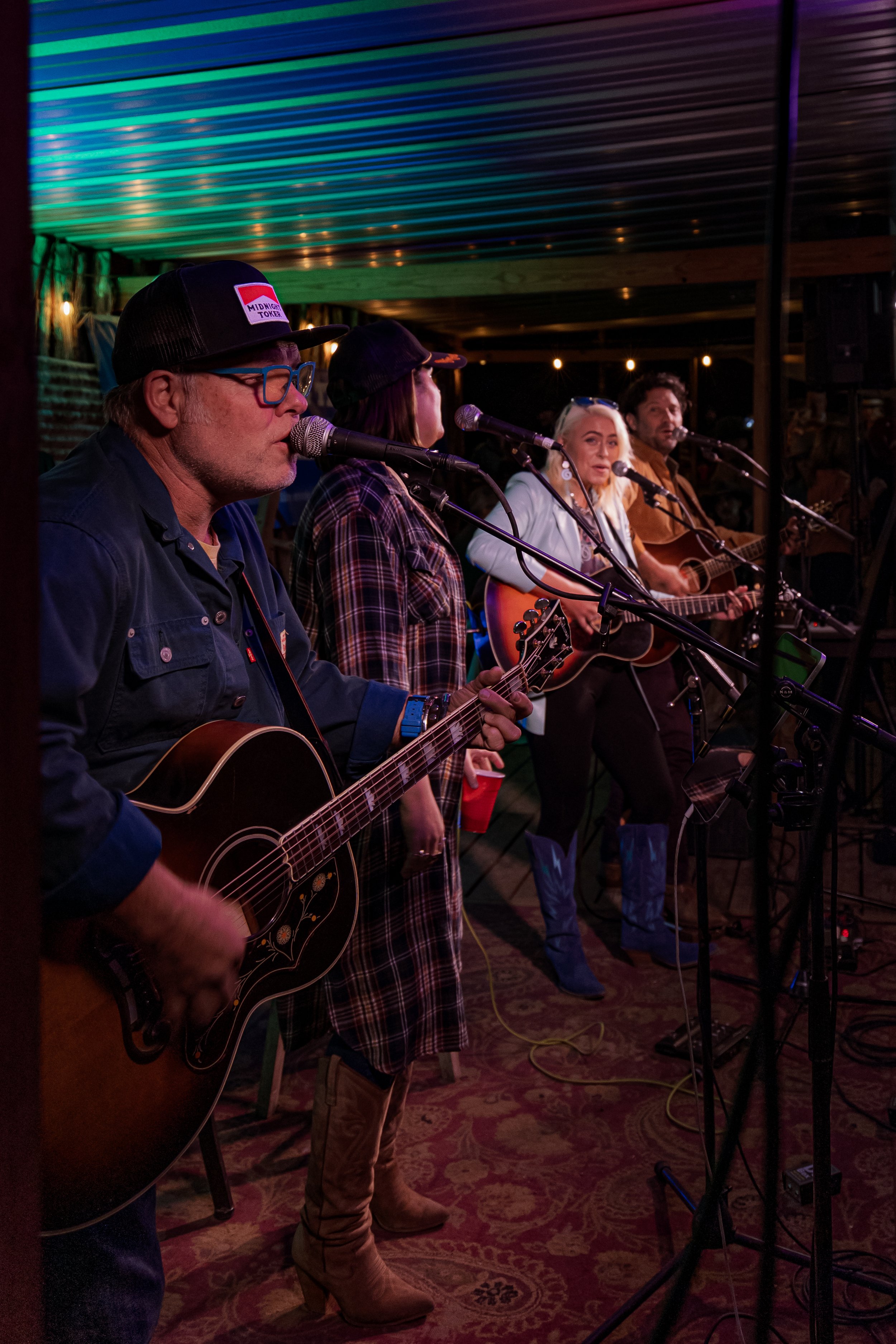 A band of five musicians performing with guitars and microphones in a dimly lit setting with colorful lighting and a patterned carpet.