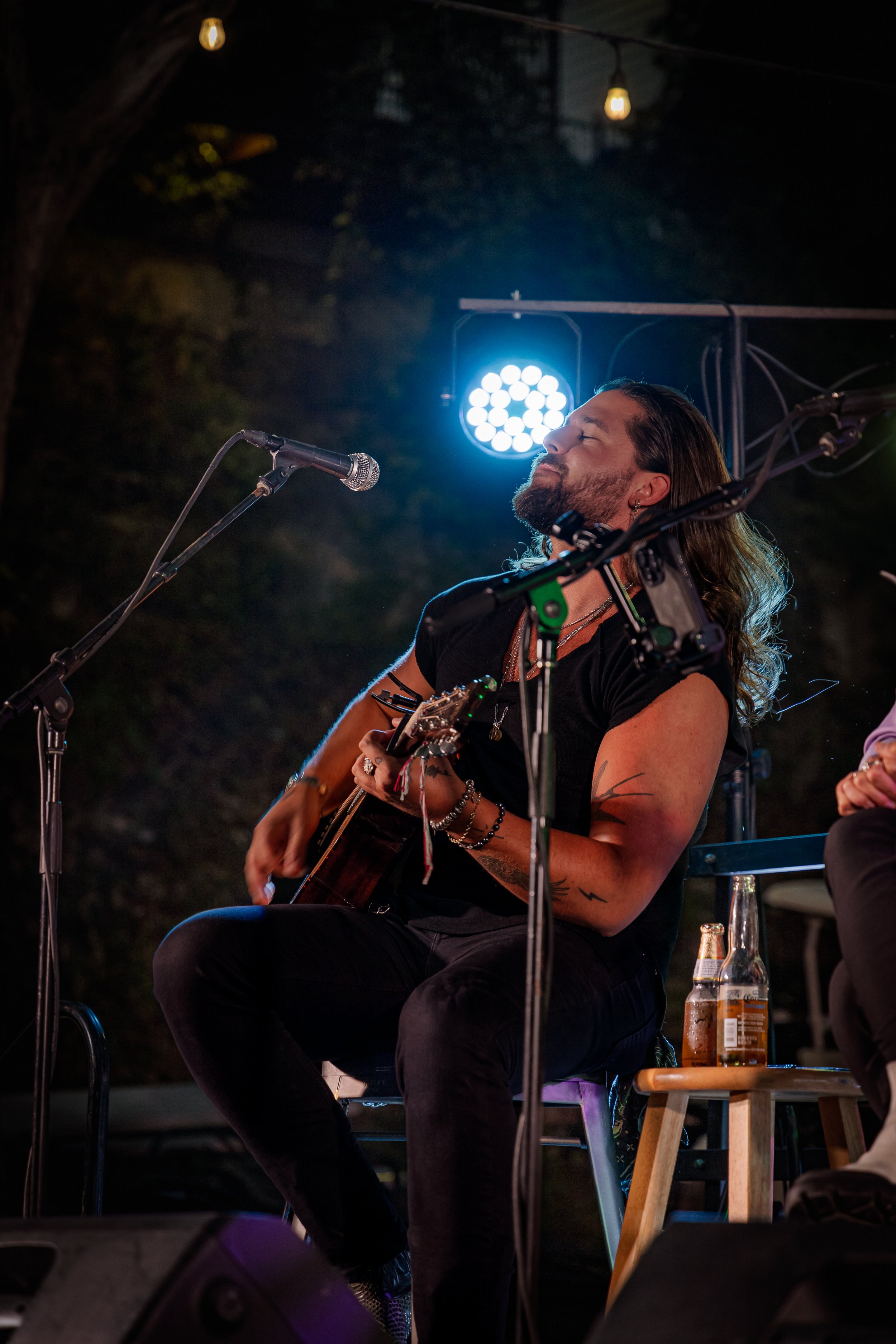 A man with long hair and a beard playing an acoustic guitar singing on stage at night, illuminated by blue stage lights, with a bottle of beer on a small wooden stool nearby.