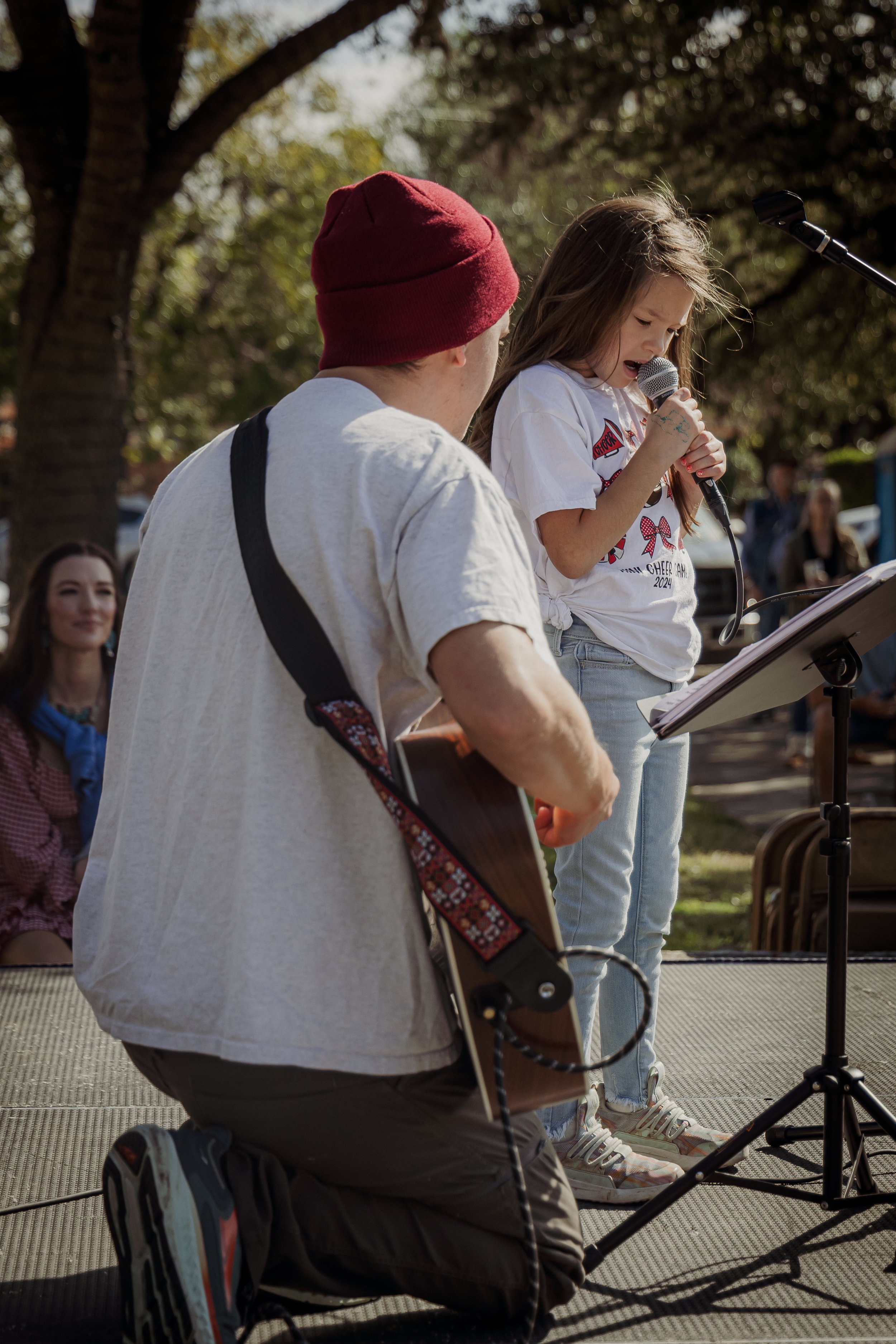 A young girl is singing into a microphone while a man is playing a guitar during an outdoor event, with audiences watching.