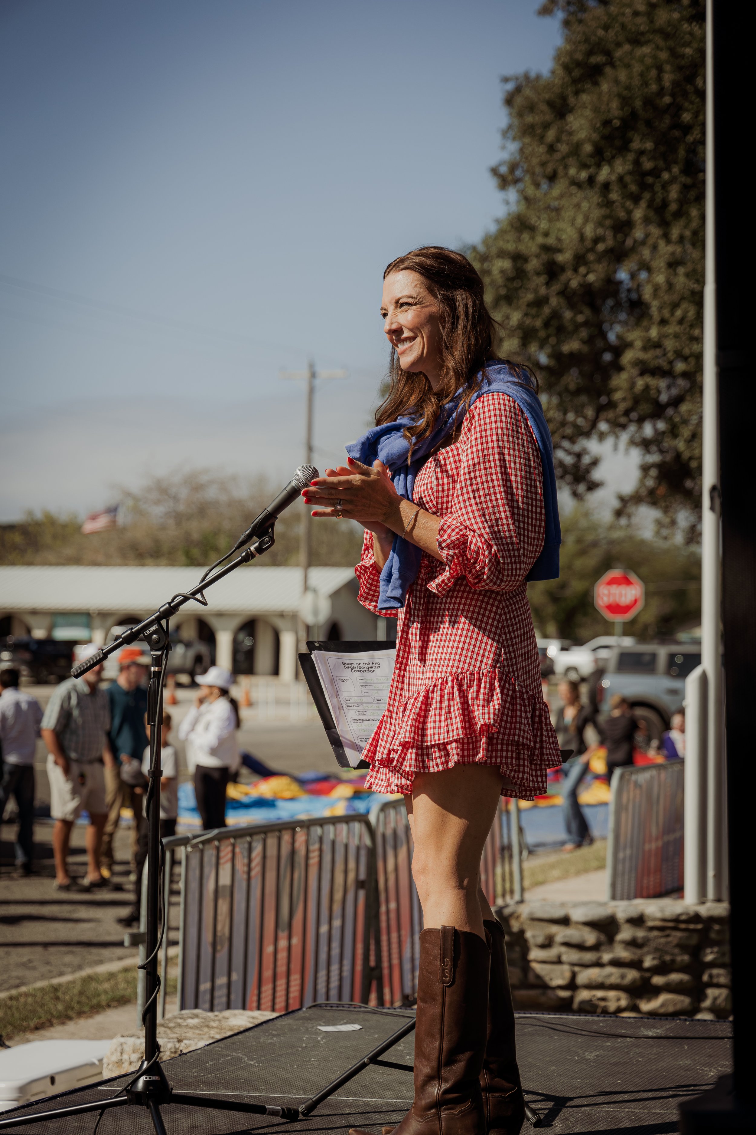 A woman in a red checkered dress with ruffles, brown boots, and a blue jacket draped over her shoulders standing at a microphone on an outdoor stage, smiling, with a crowd and a stop sign in the background.