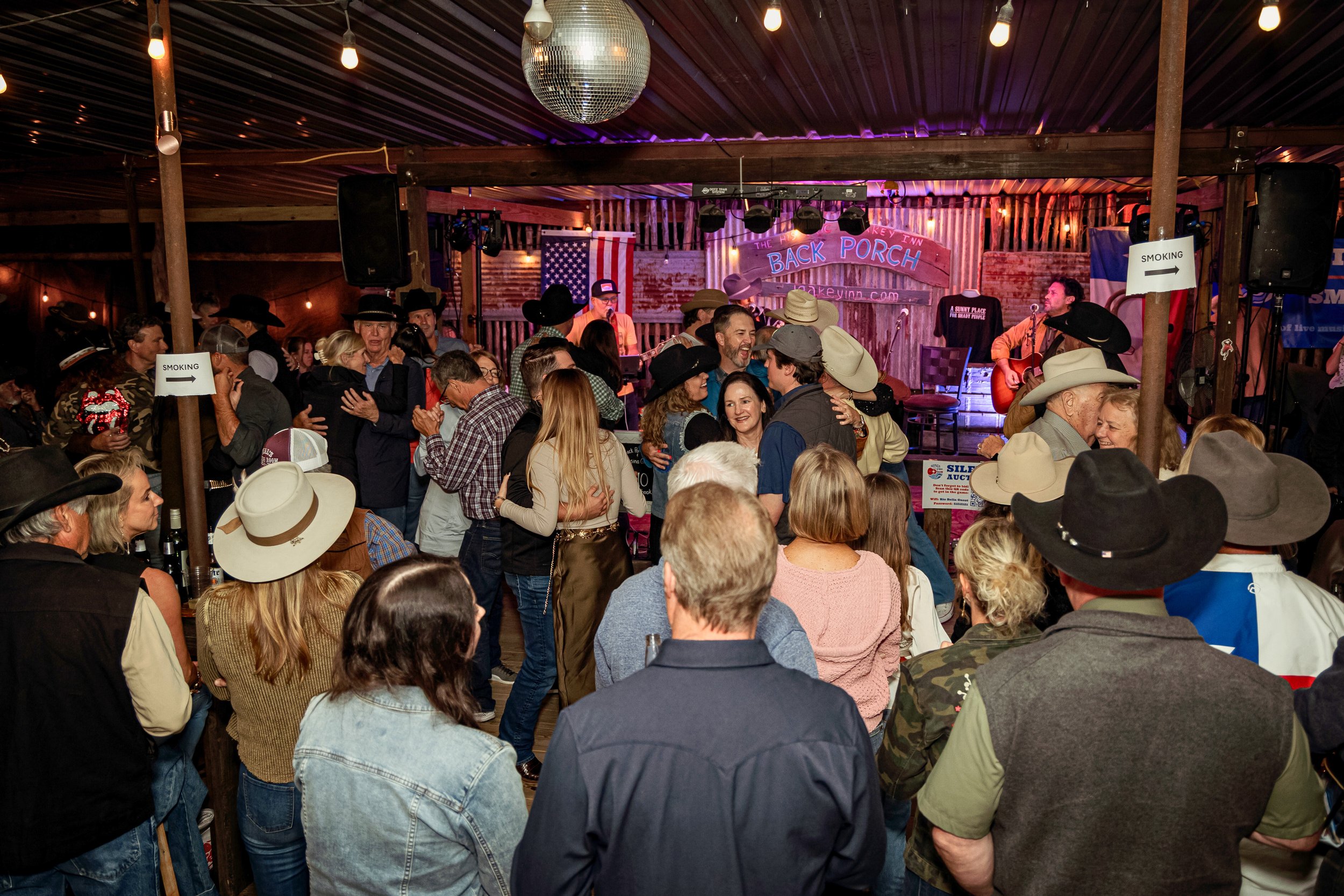 Crowd dancing and socializing at a country-themed event with musicians on stage, cowboy hats, and rustic wooden decor.
