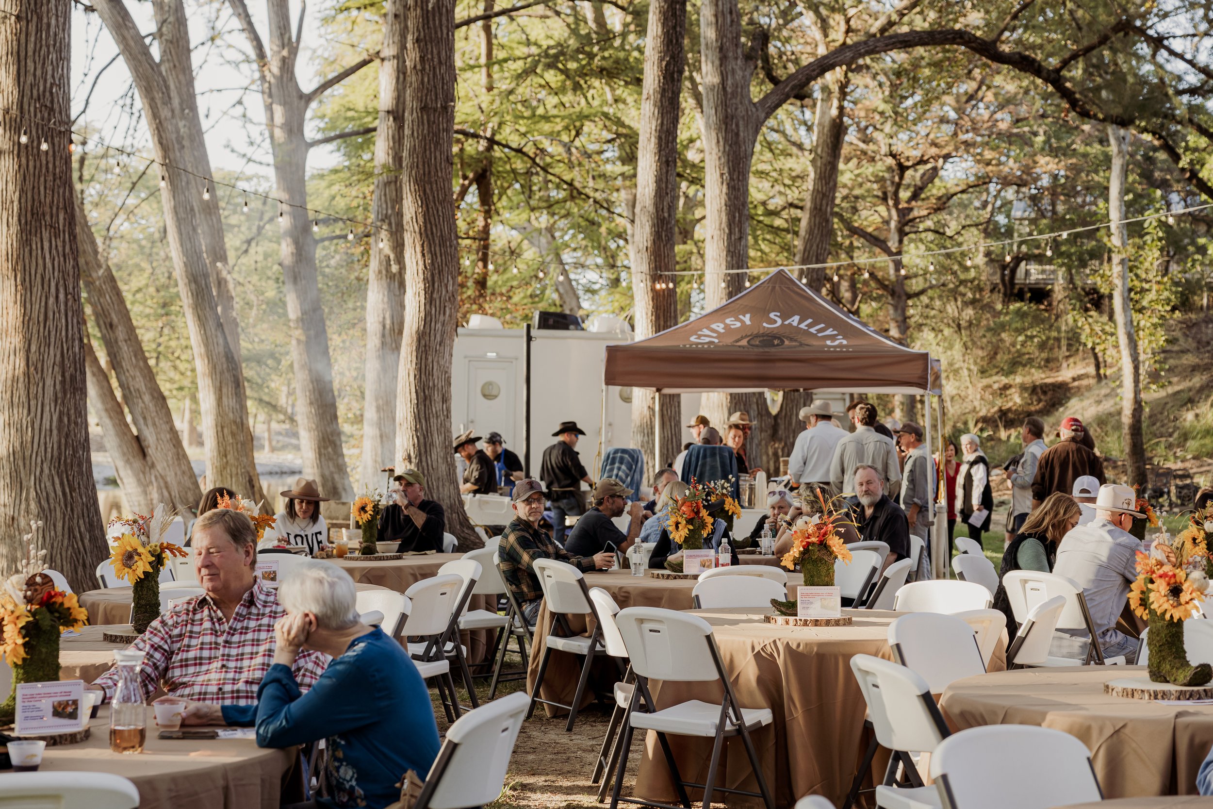People enjoying an outdoor gathering with tables decorated with sunflowers and fall-themed centerpieces under tall trees.