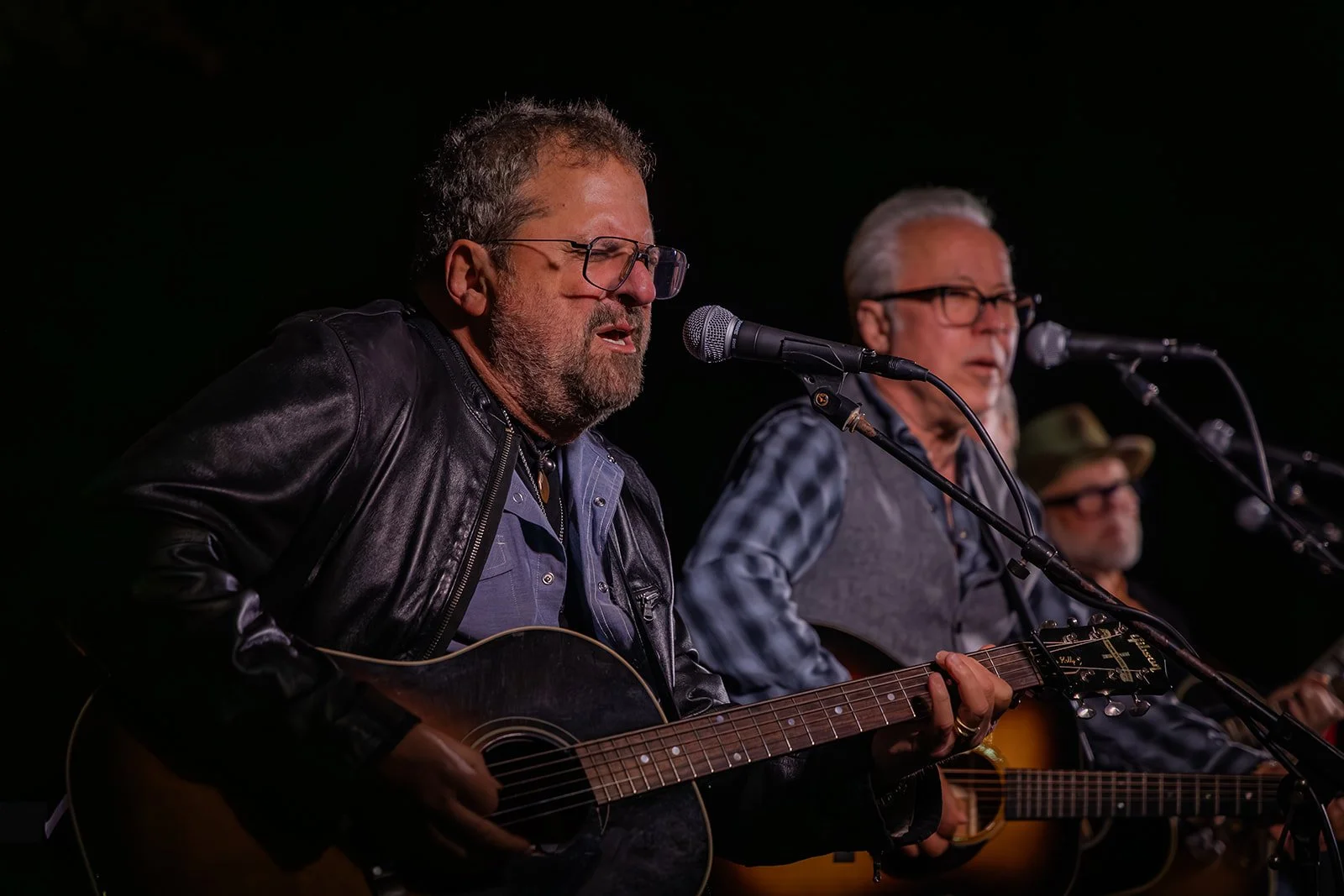 Three men performing with guitars on stage, singing into microphones, with dark background.