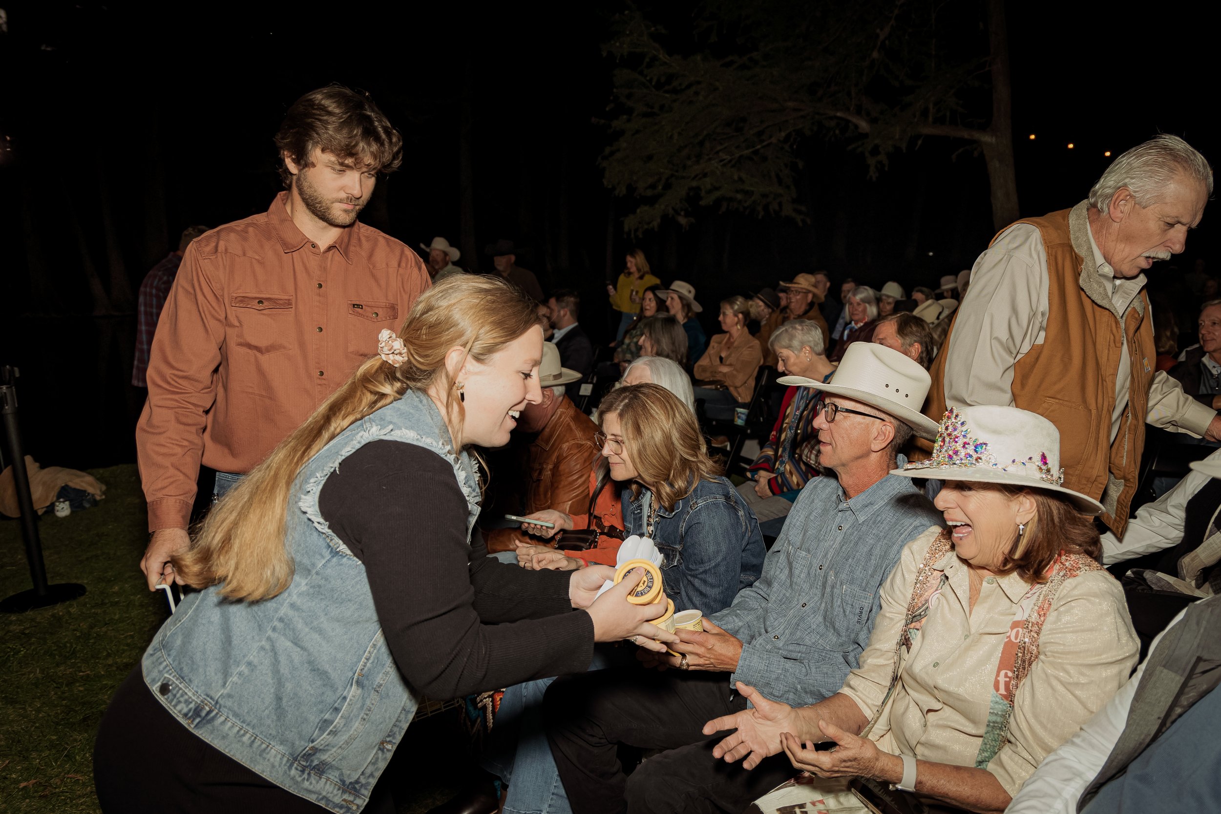 A young woman is happily handing out small items to seated older people at an outdoor event during the evening, with many other attendees in the background. The scene suggests a friendly, festive gathering.