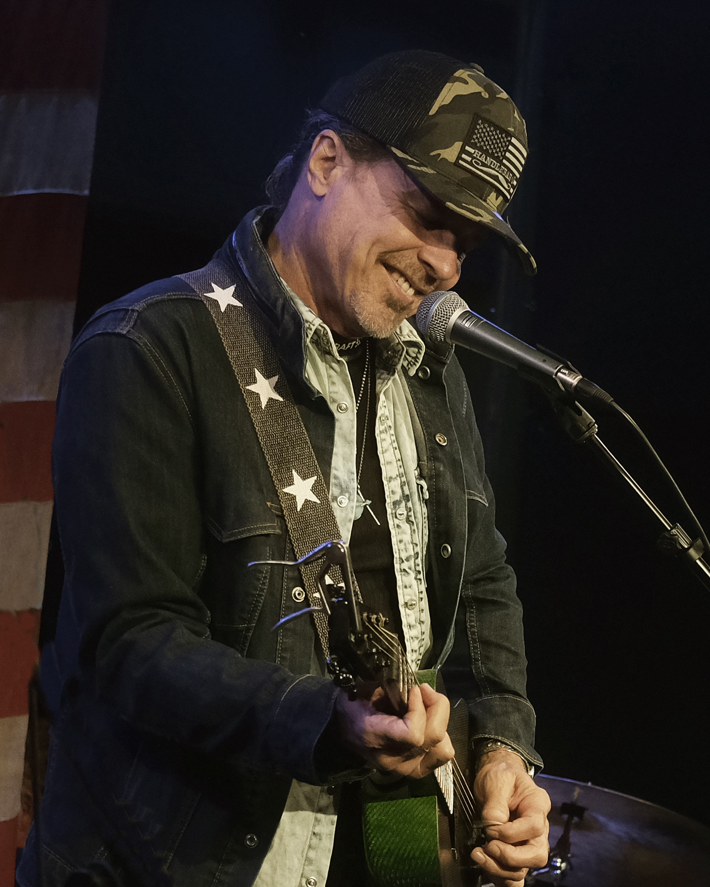 Male musician with closed eyes singing into a microphone while playing an acoustic guitar, wearing a black hat and black shirt, against a warm yellow-orange background.