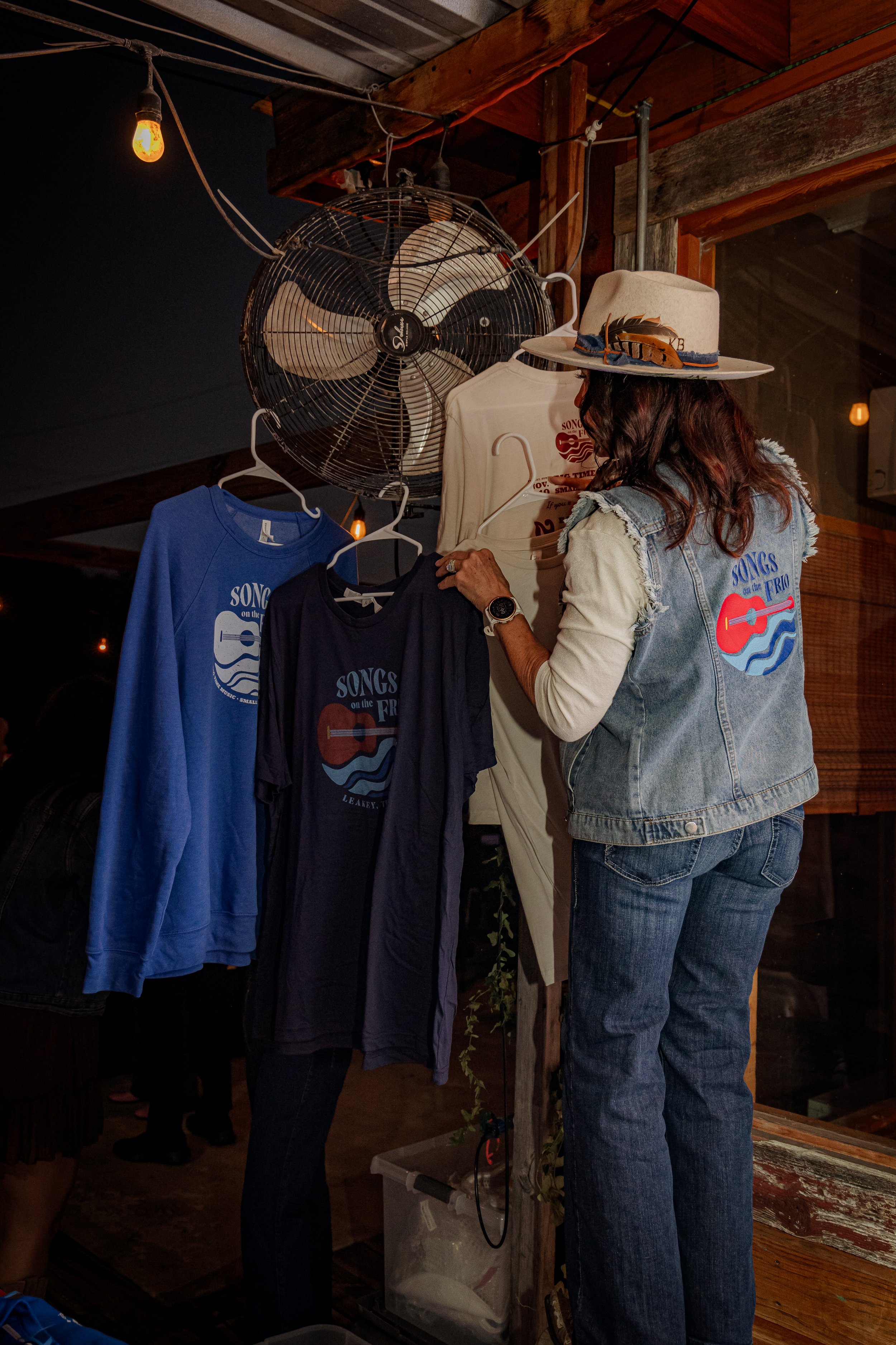 A woman with a watch on her wrist is browsing t-shirts with a musical theme that say "Songs on the Frio." The t-shirts are hanging on hangers from a piece of display equipment with a fan above. The woman is wearing a denim vest, jeans, and a hat with
