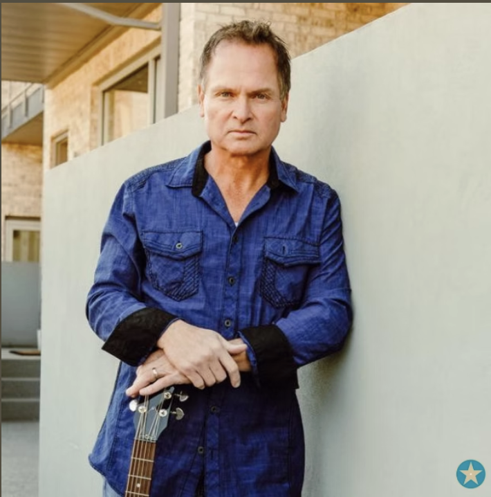 A middle-aged man with short brown hair, wearing a blue denim shirt with black cuffs, standing outdoors against a concrete wall. He is holding a guitar with his right hand and has a serious expression.