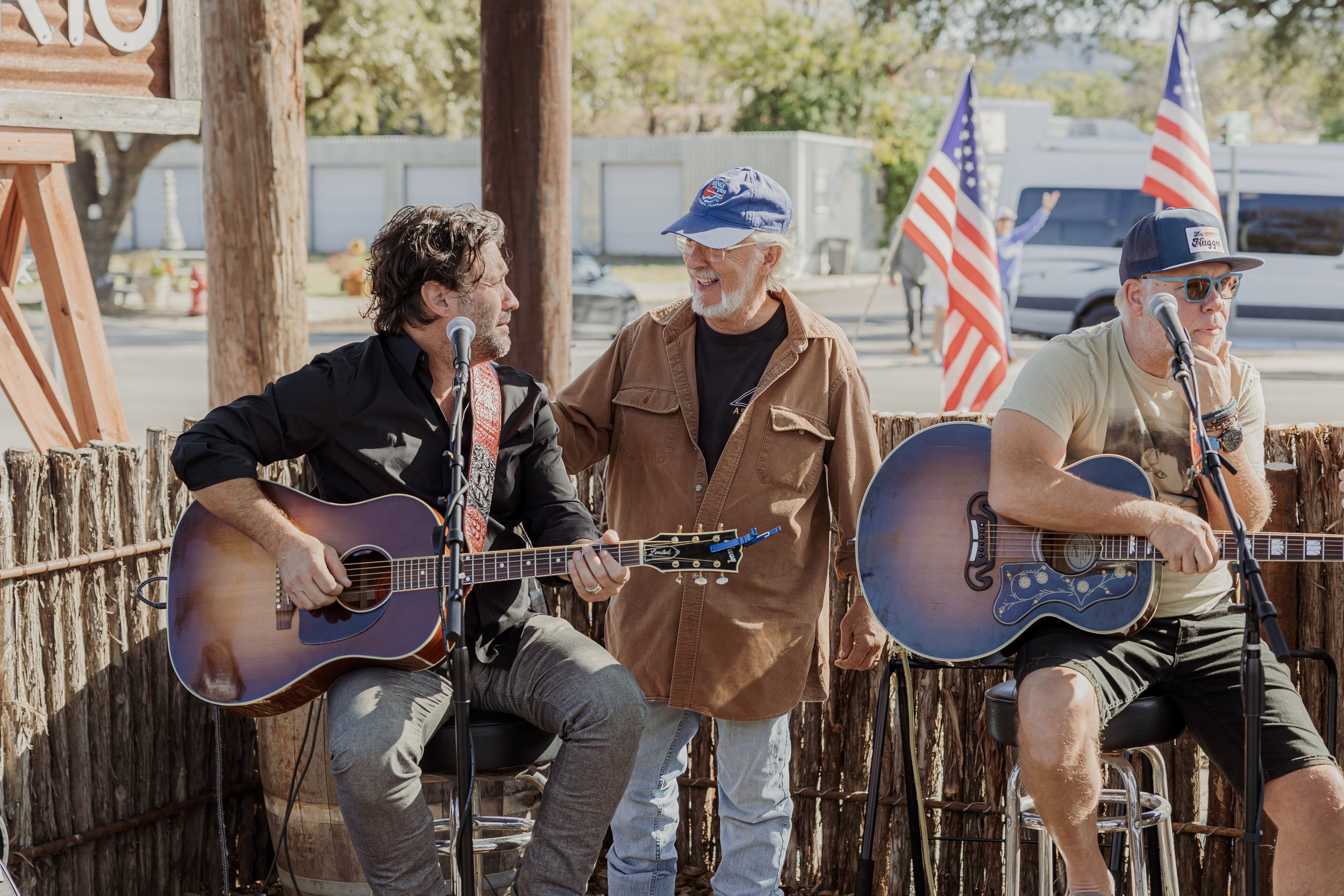 Two men playing guitars and a third man standing between them, smiling and talking at an outdoor event, with American flags in the background.