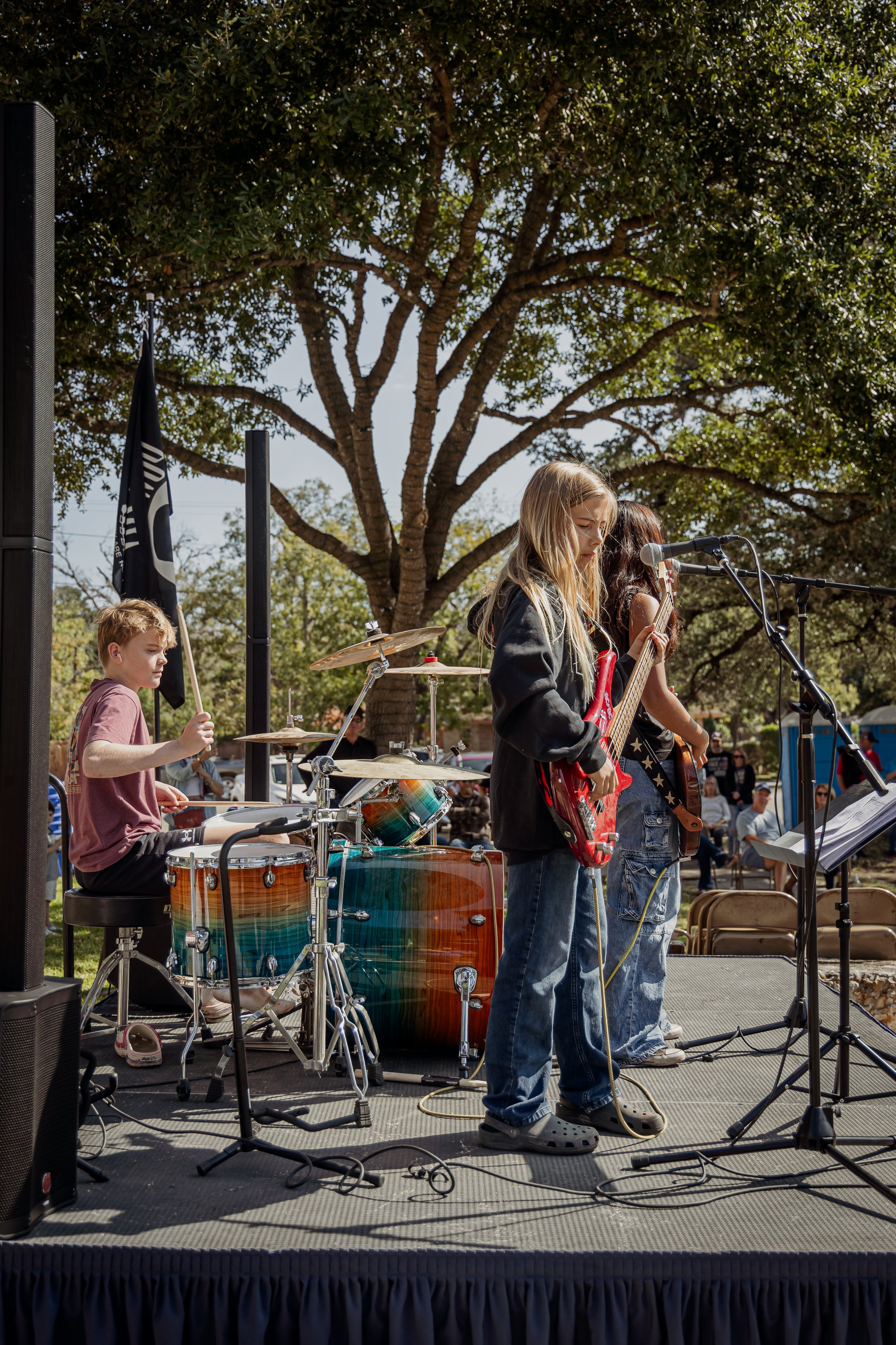 Young children performing in a band on an outdoor stage, with one playing drums, another singing into a microphone, and a third playing an electric guitar under a large tree.