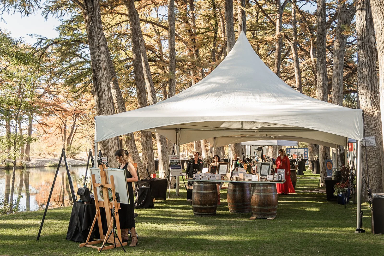 Outdoor art fair under a white canopy tent surrounded by tall trees near a body of water, with several people viewing and selling artwork on display.