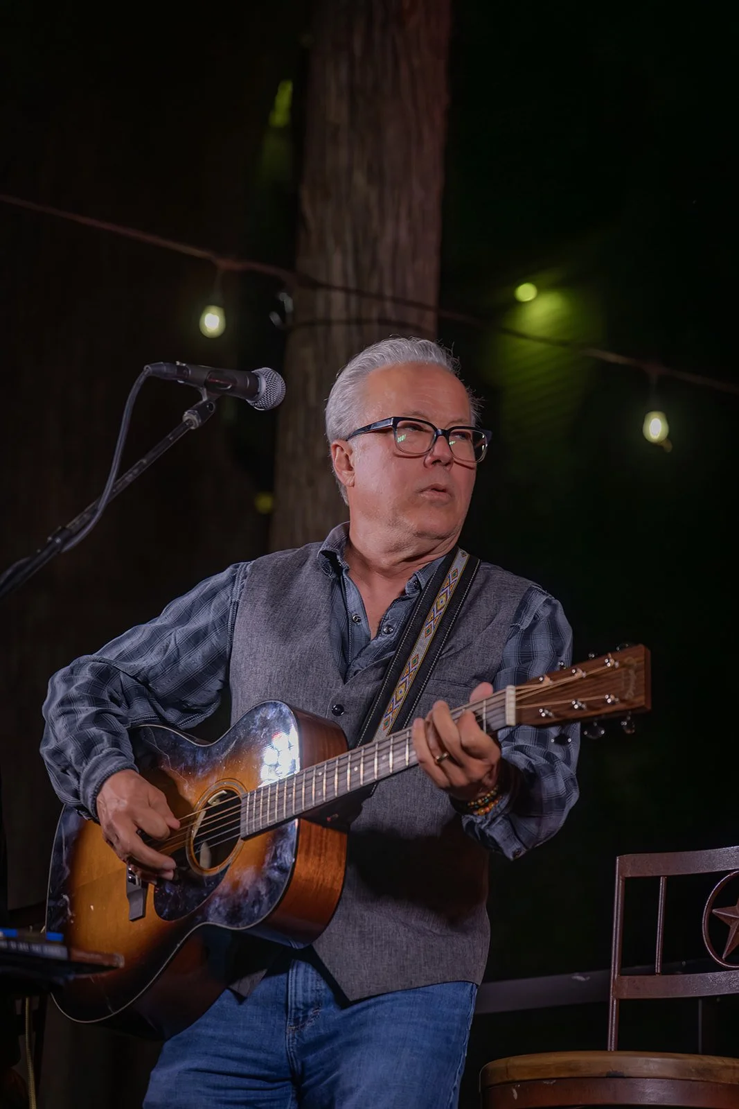 An older man with glasses playing an acoustic guitar on stage, with a microphone nearby and string lights in the background.