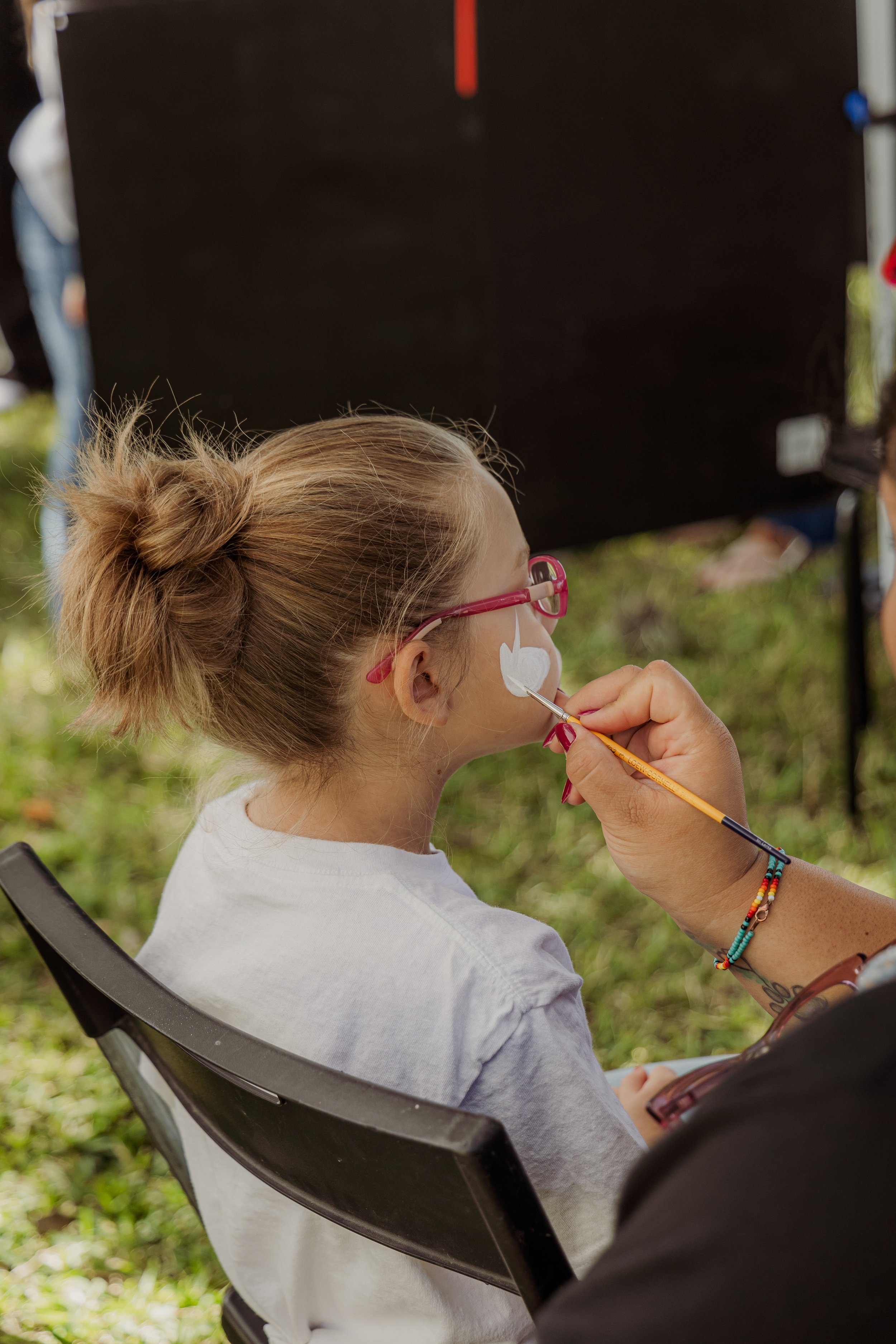 A young girl with blonde hair tied in a messy bun, wearing pink glasses, is getting her face painted with a white heart on her face outdoors. An artist's hand with bracelets is holding a paintbrush near her cheek.