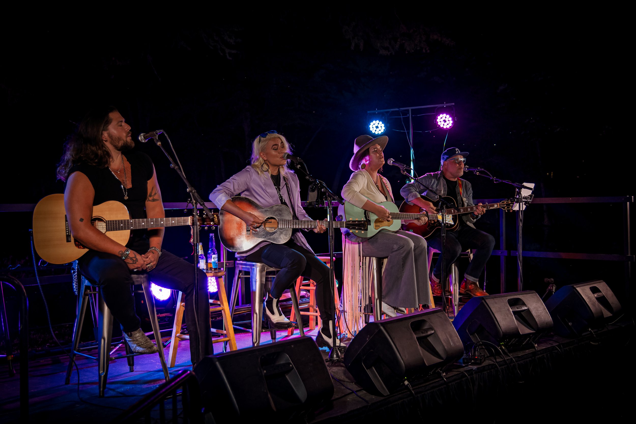 Four musicians sitting on stage with guitars, performing at night, illuminated by colorful stage lights.