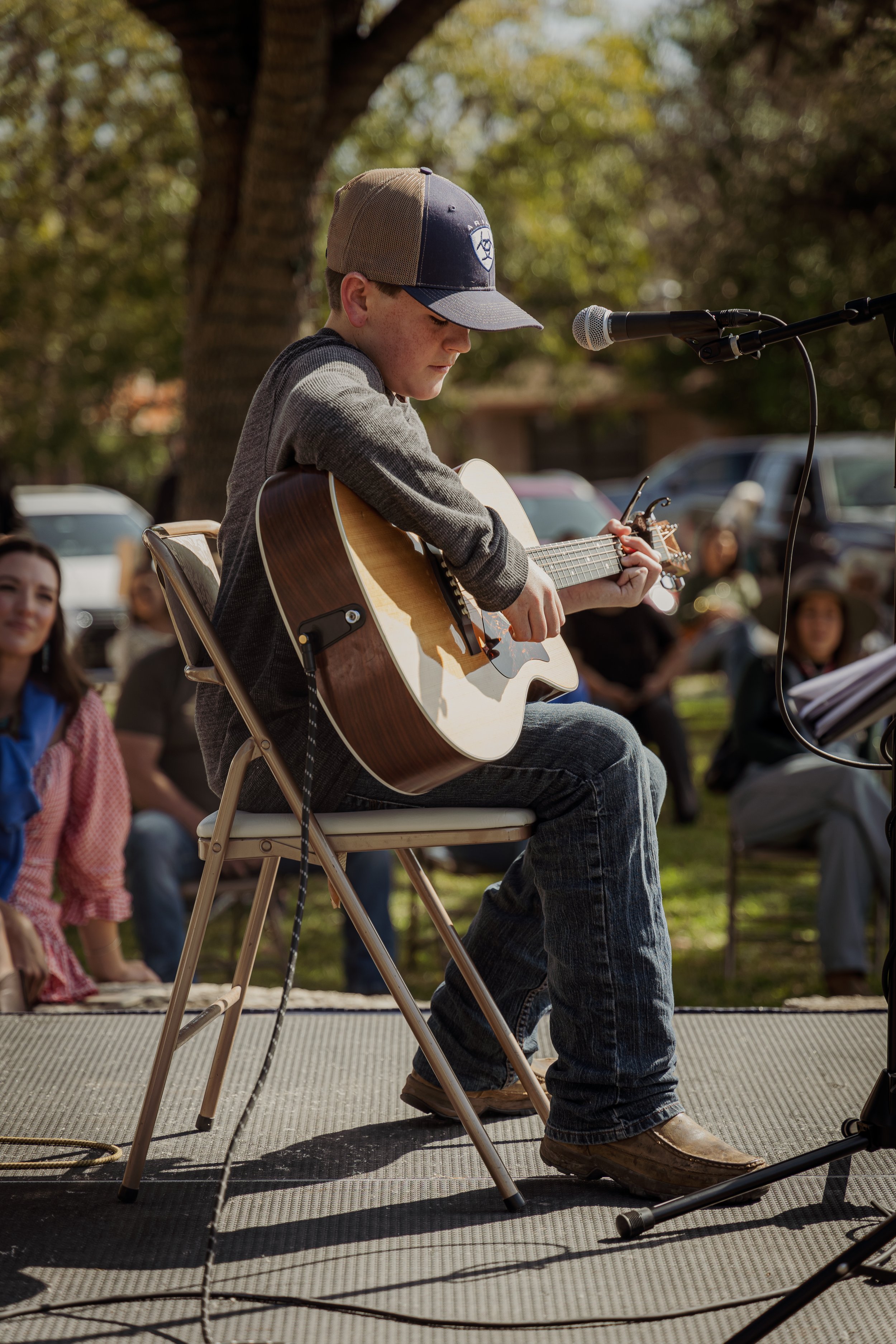 A young boy sitting on a chair playing an acoustic guitar outdoors during a music performance, with an audience seated behind him and a large tree overhead.