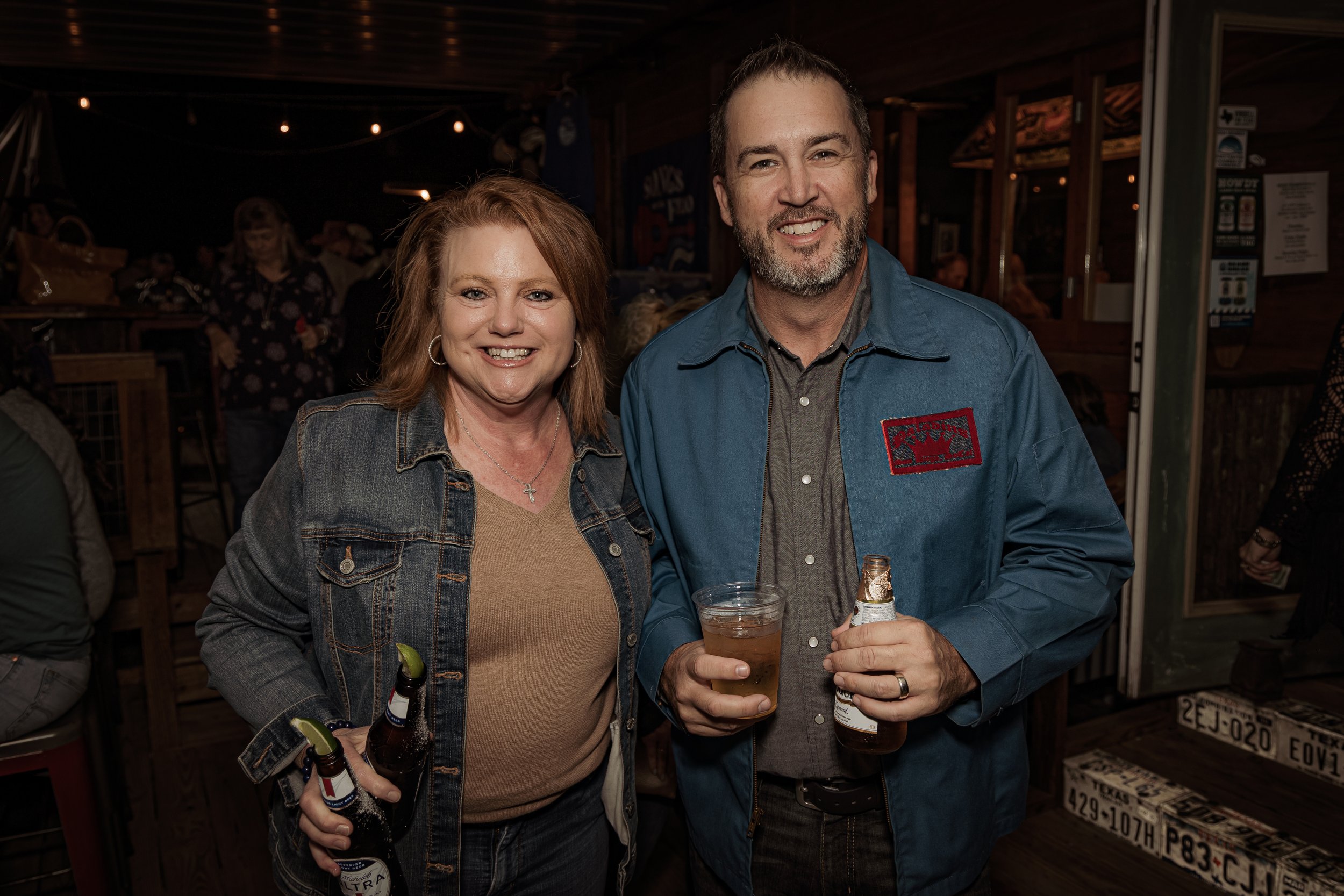 A man and woman smiling and holding drinks at a bar; woman holding two bottles, man holding a glass of beer and a bottle, in a dimly lit rustic setting.