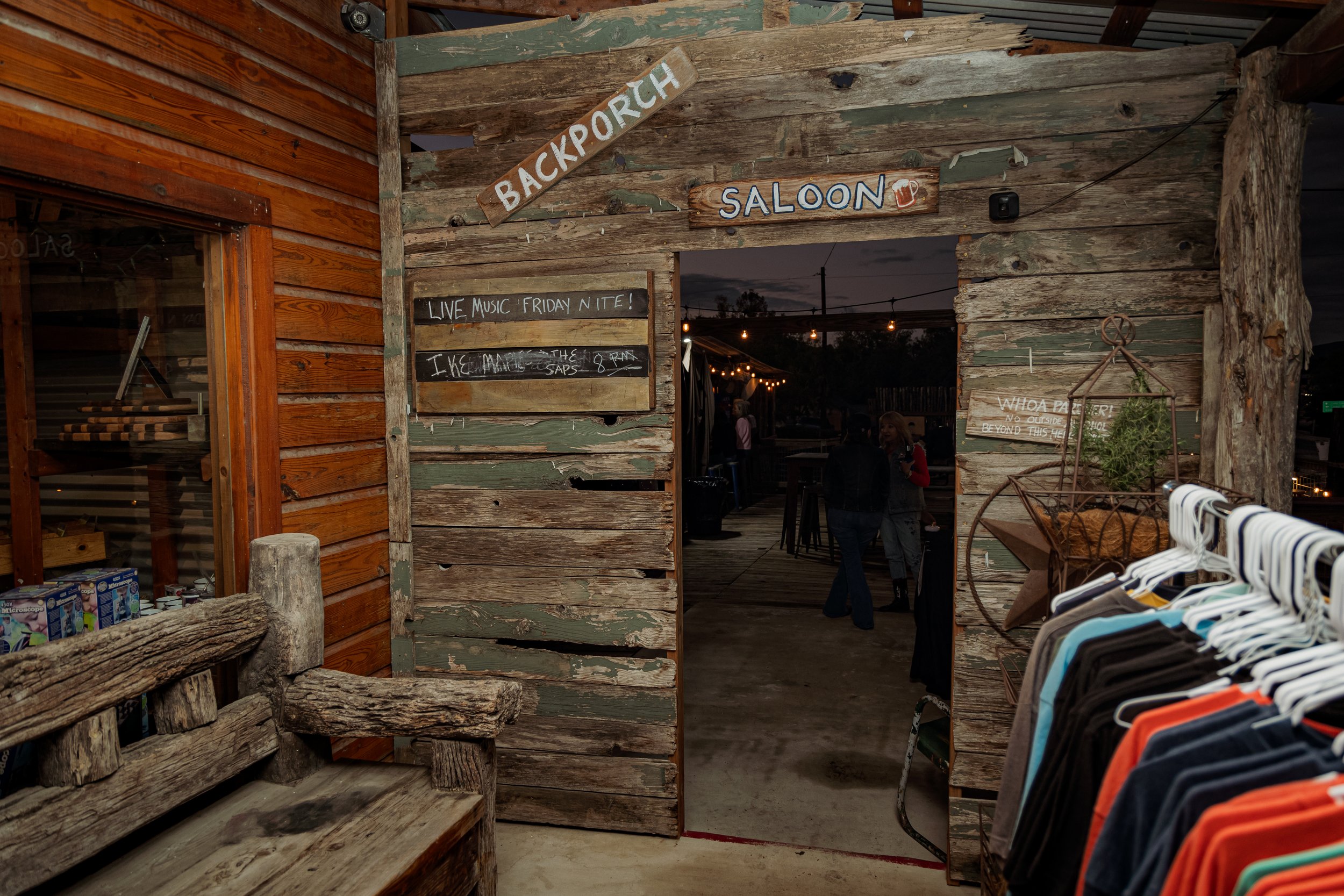 Rustic wooden entrance to a bar or pub, with signs reading 'BACKPORCH' and 'SALOOK,' entering into an outdoor social area. Inside, there's a string of lights, a chalkboard sign advertising live music on Friday night, and several people socializing ar