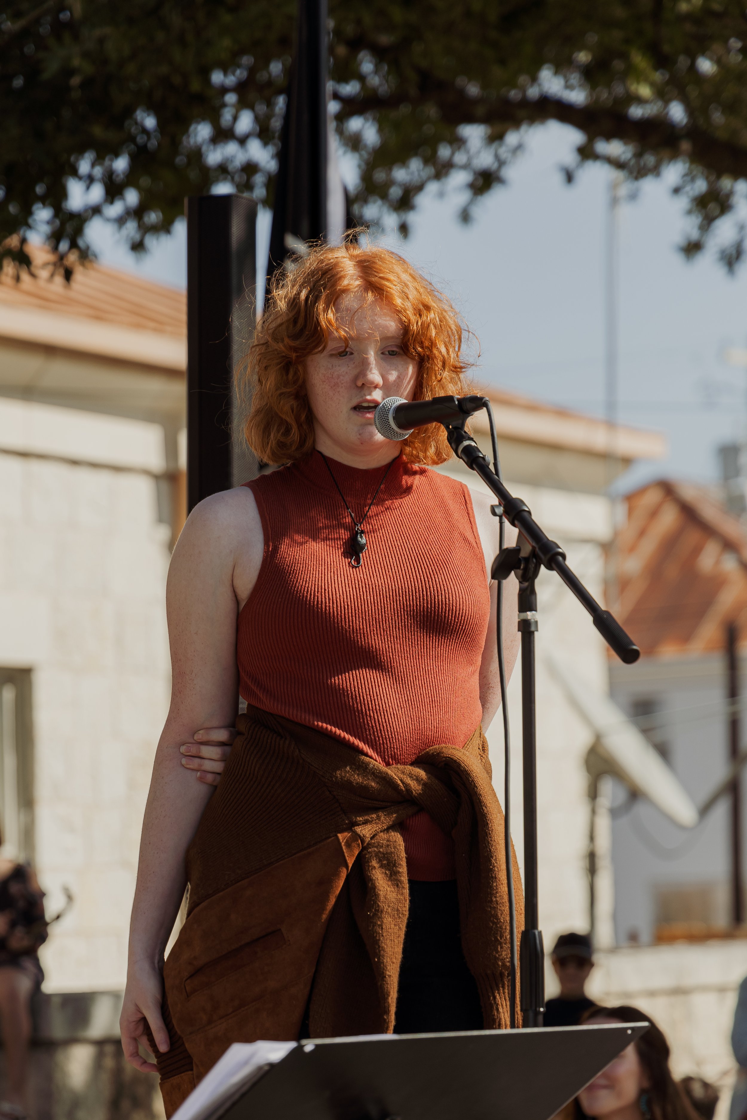 A young woman with curly red hair, wearing a sleeveless rust-colored top and a brown jacket tied around her waist, stands outdoors in front of a microphone, speaking or singing. She appears focused, with a tree and buildings in the background.
