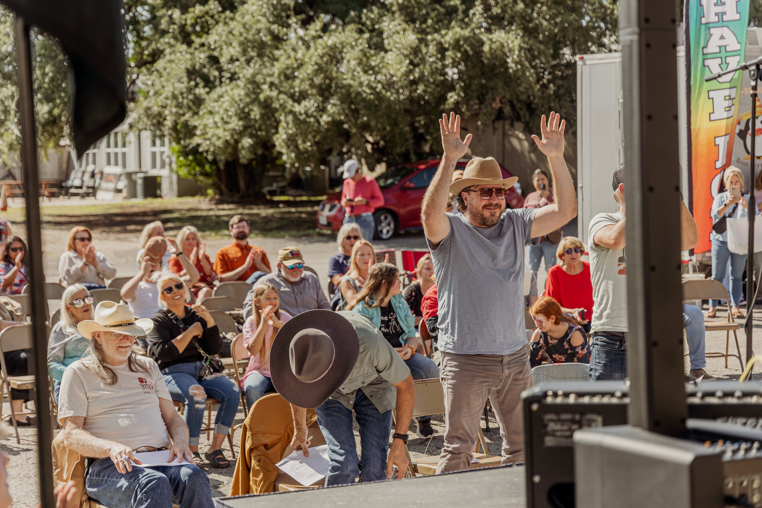 Man wearing a tan hat, sunglasses, and a gray t-shirt with his hands raised, standing in front of an outdoor audience at a daytime event.