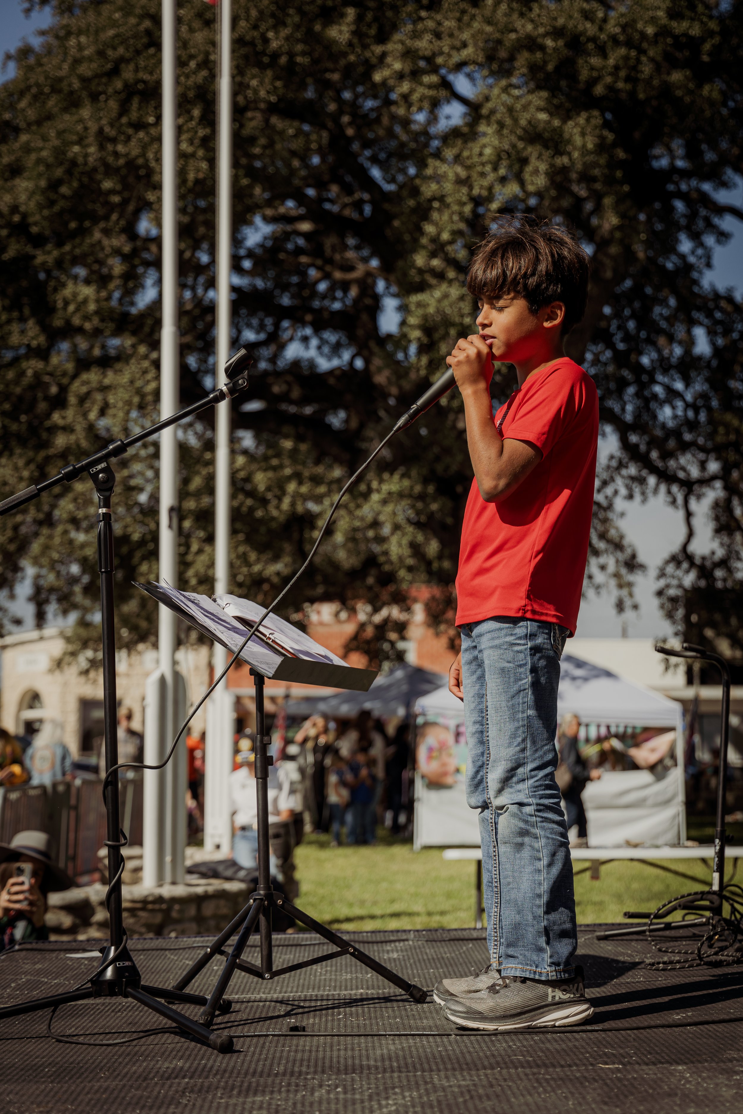 A young boy in a red shirt and jeans singing into a microphone on an outdoor stage during a daytime event, with a crowd and trees in the background.
