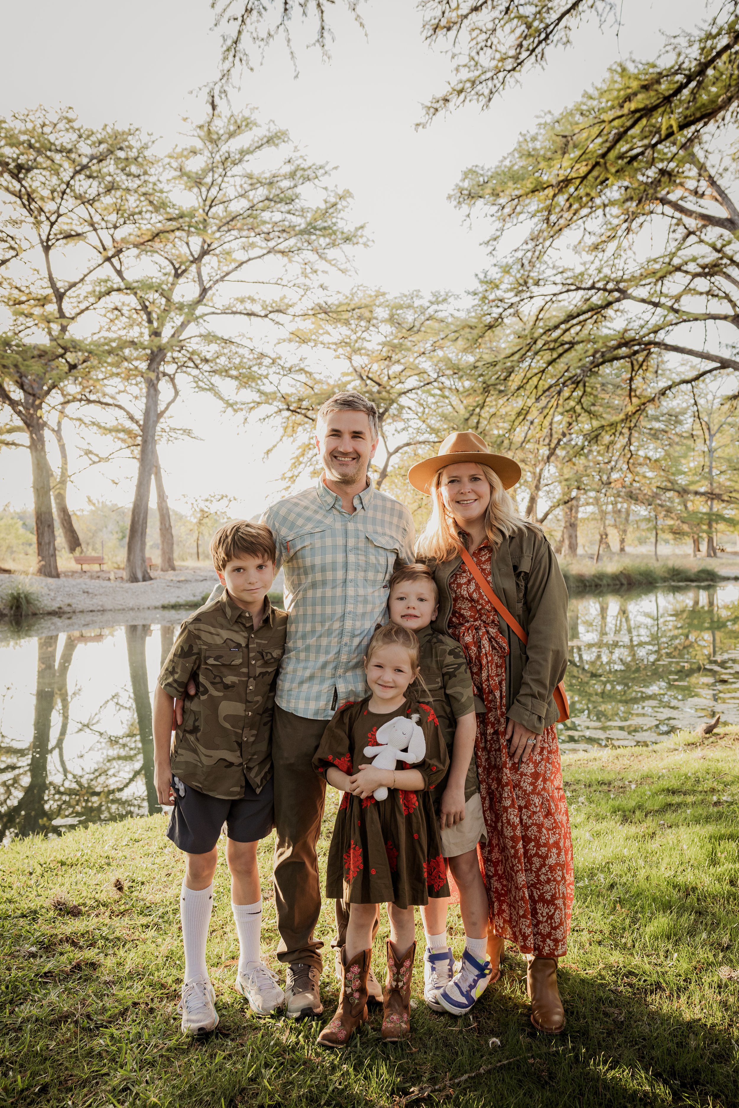 Family of six posing outdoors near a pond with trees in the background, all smiling.