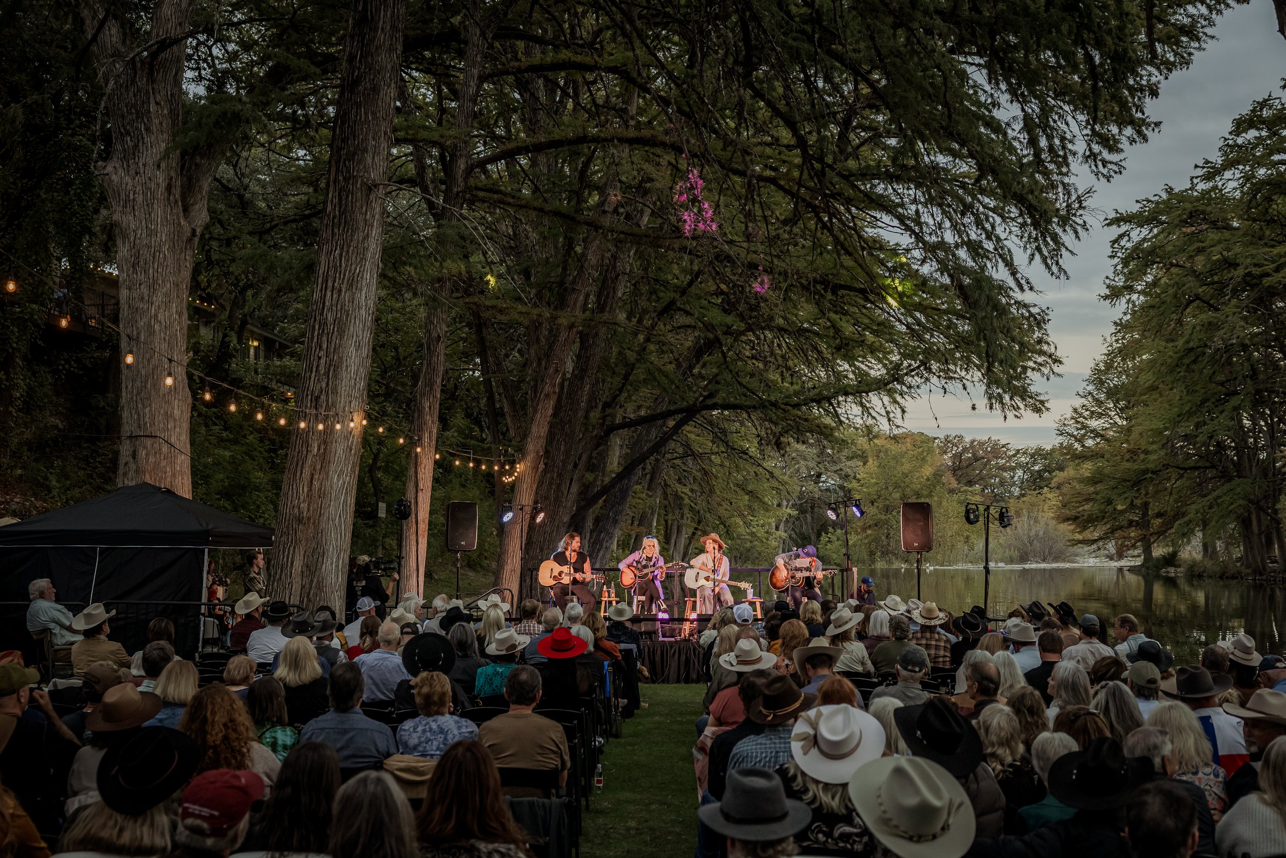 An outdoor concert taking place in a wooded area by a lake, with a large audience seated and watching a band perform on a stage surrounded by tall trees.