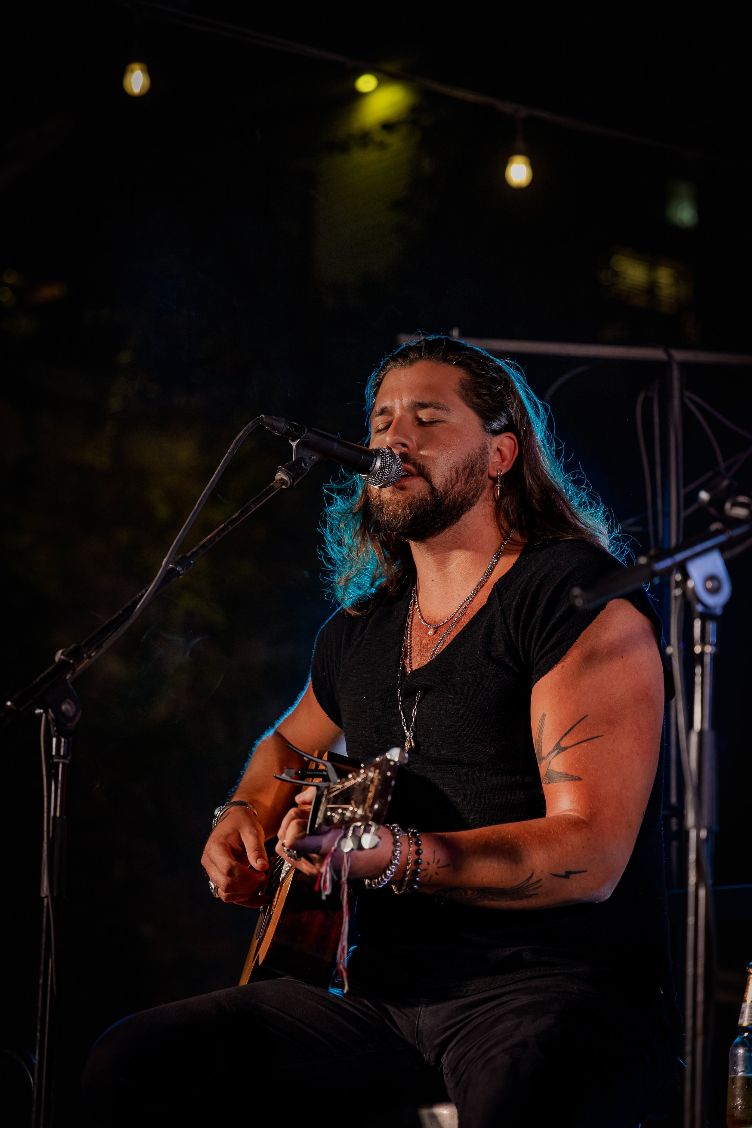 Male musician with long hair, tattoos, and jewelry, singing into a microphone while playing an acoustic guitar at night.