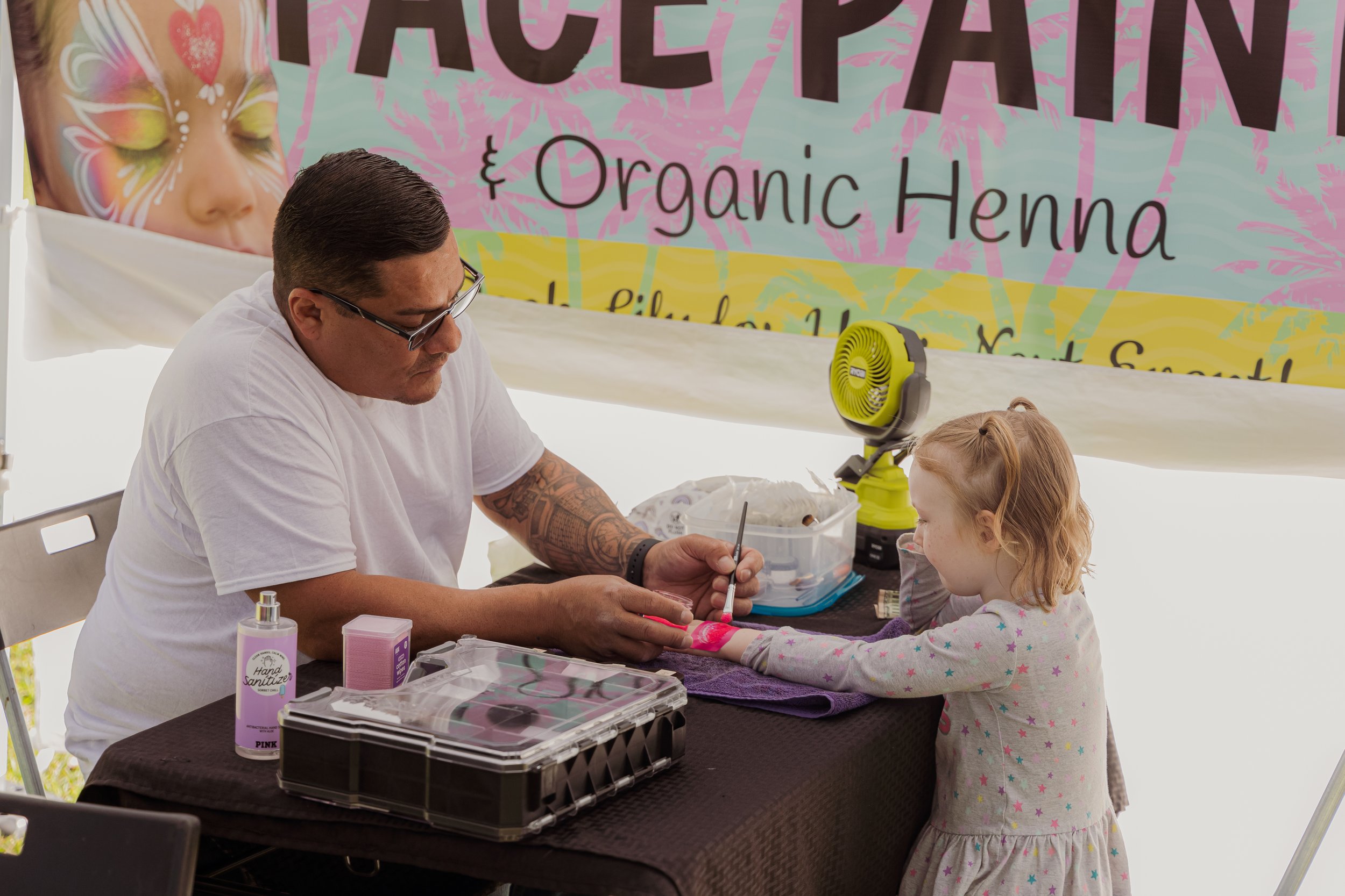 A man tattooing a young girl’s wrist at a booth with a sign that reads 'Organic Henna' in the background.