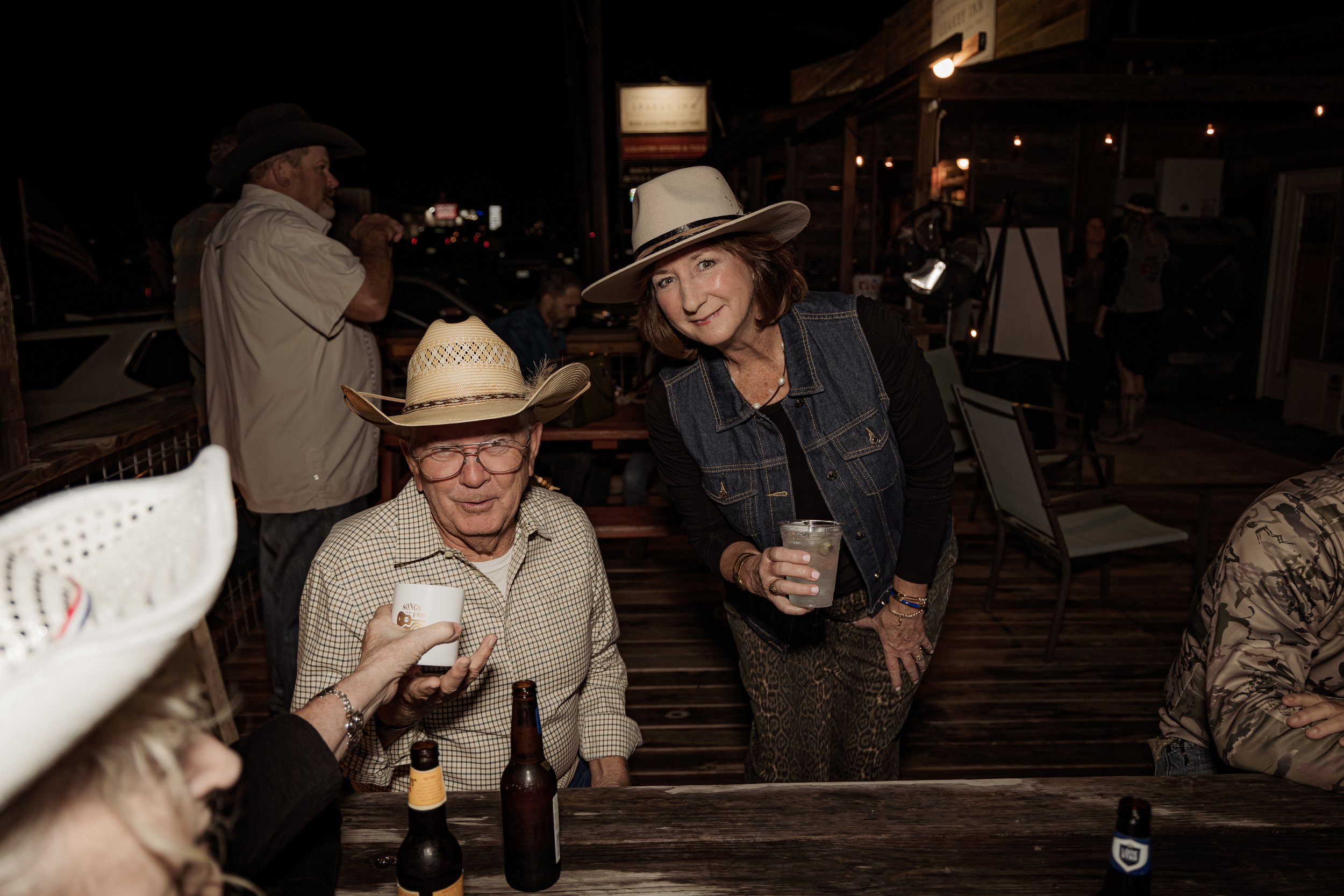 Group of people at a western-themed bar or restaurant, wearing cowboy hats, with drinks and beer bottles on the table, in an outdoor or semi-outdoor setting during the evening.