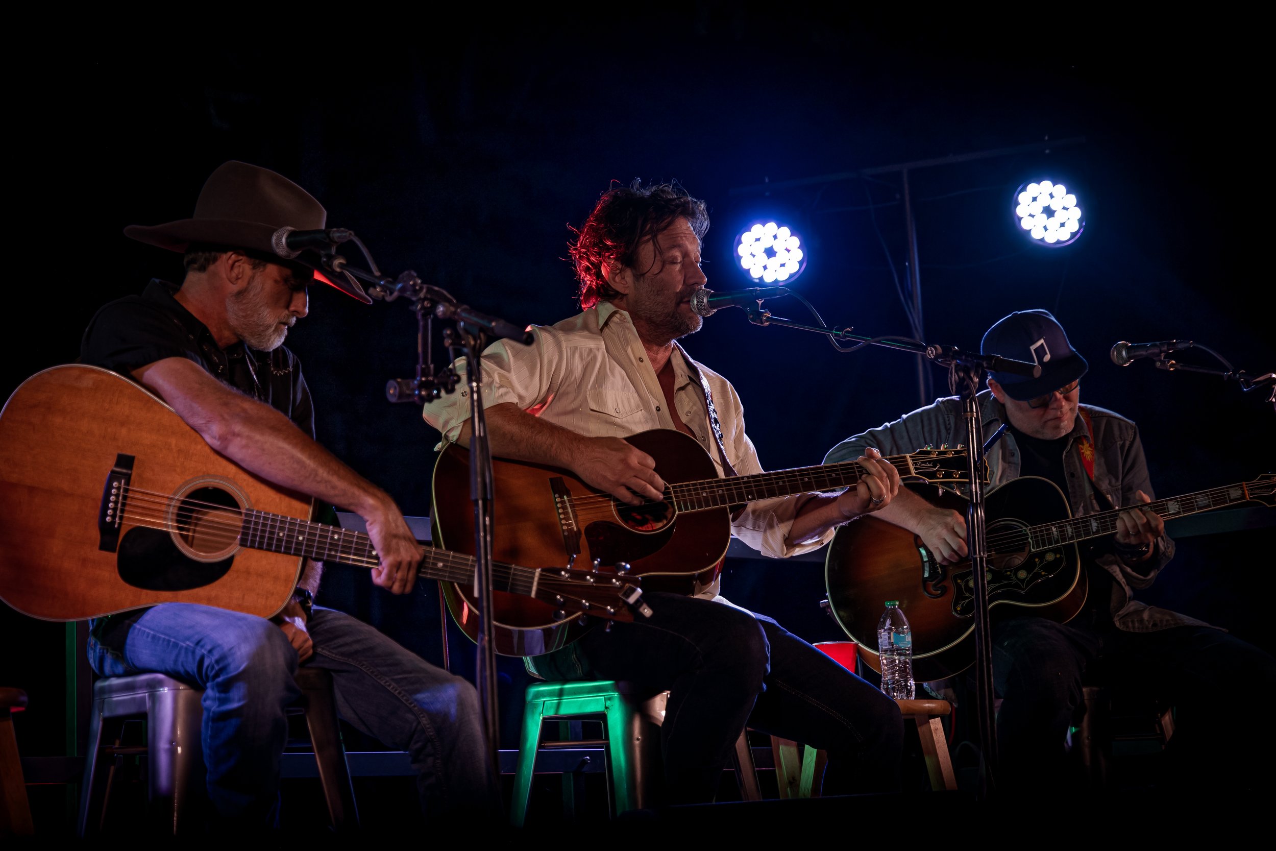 Three male musicians playing acoustic guitars on stage under blue and white stage lights.