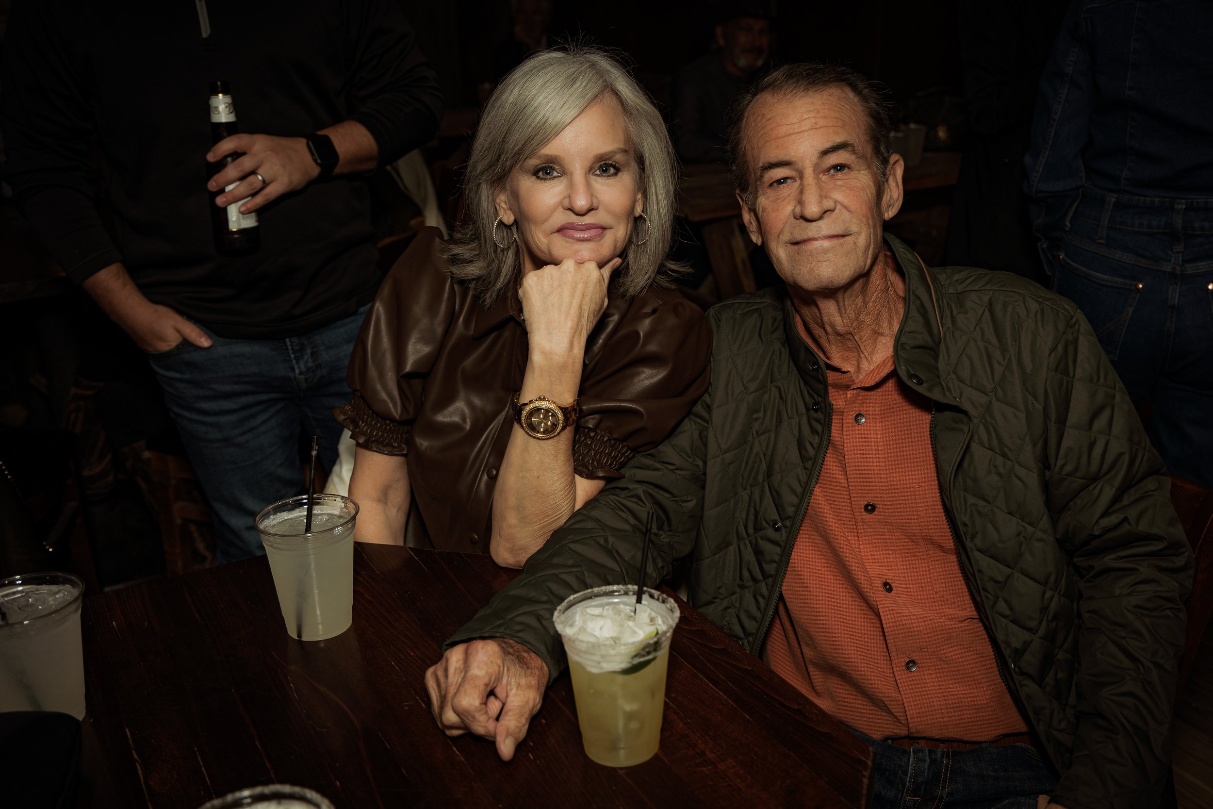 A woman and an older man sitting at a table with drinks, posing for a photo in a dimly lit setting, possibly a bar or restaurant.