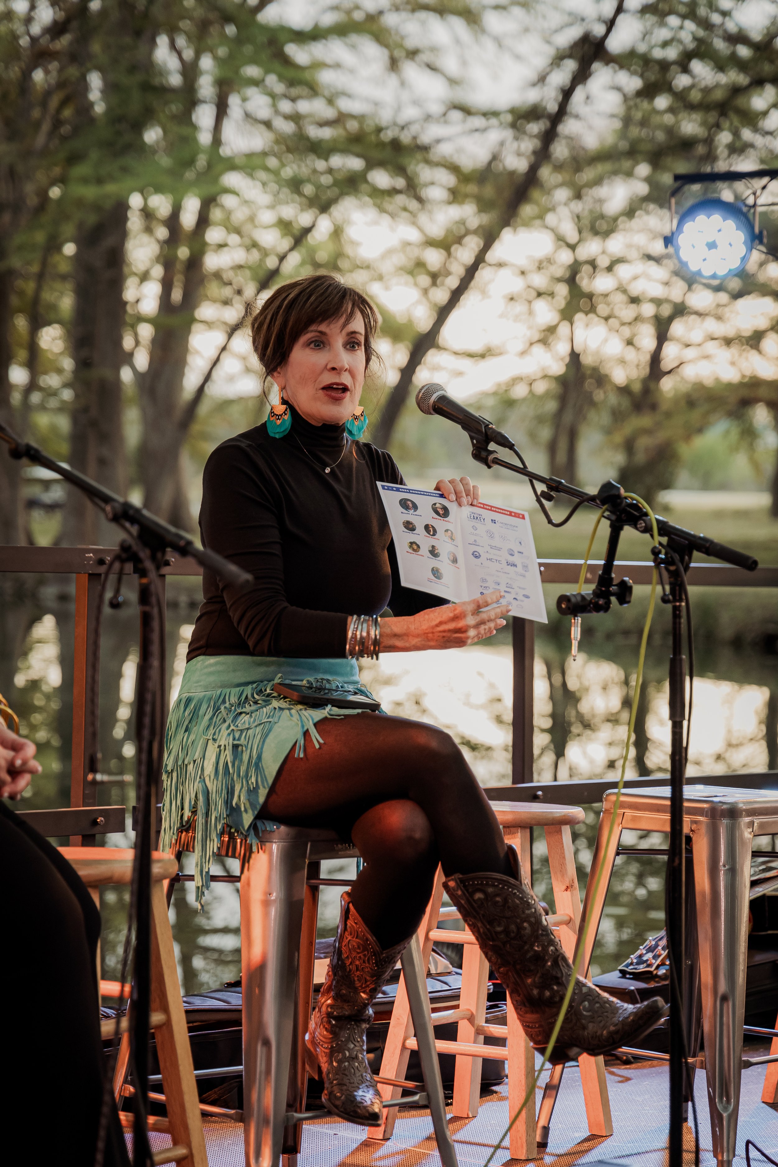 A woman with short brown hair, wearing a black top, turquoise fringed skirt, black tights, and cowboy boots, is sitting on a stool outdoors. She is holding a brochure or flyer and speaking into a microphone, with trees and a body of water in the back