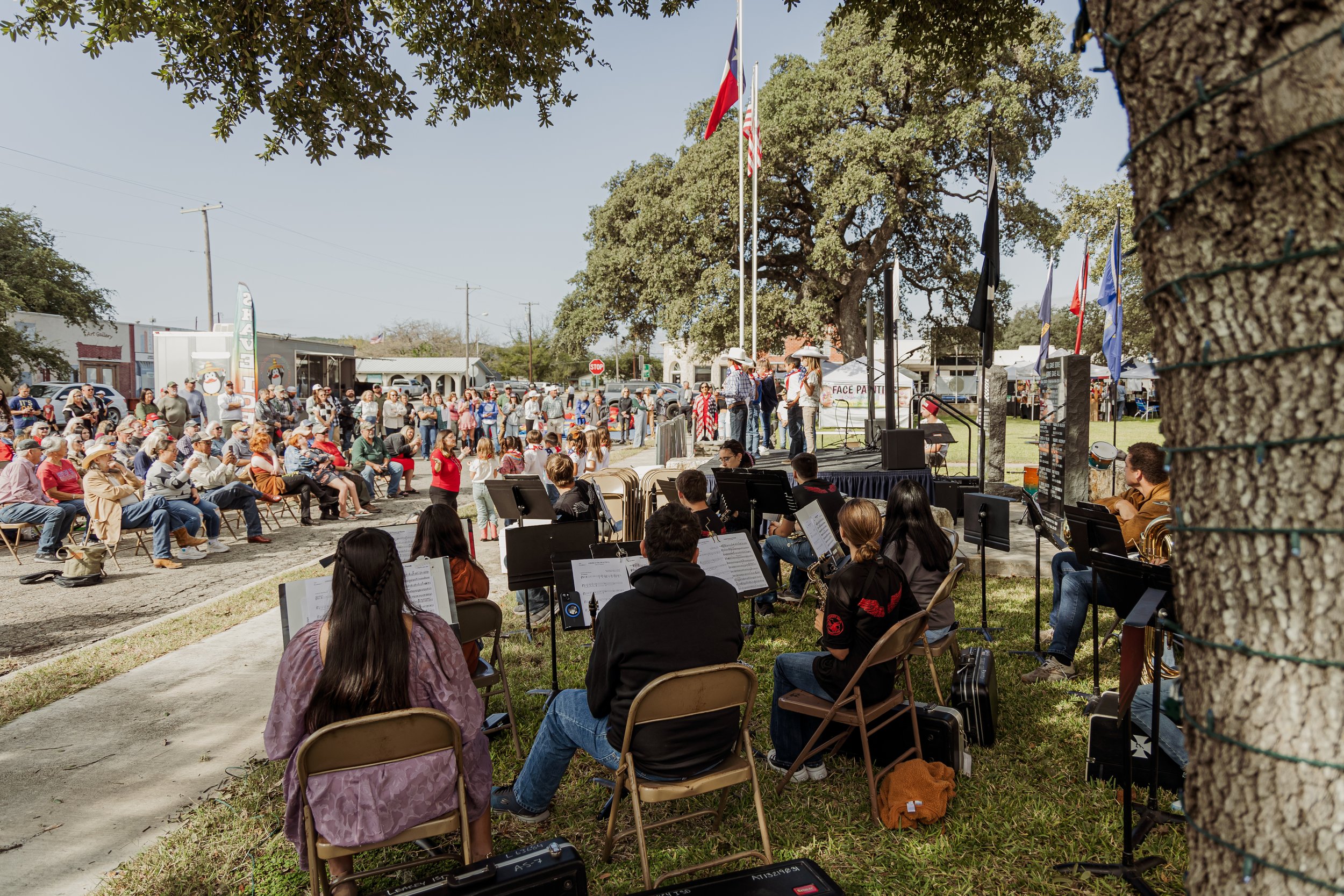 A community outdoor event with a stage featuring singers, a seated orchestra, and an audience gathered under a large tree with flags.