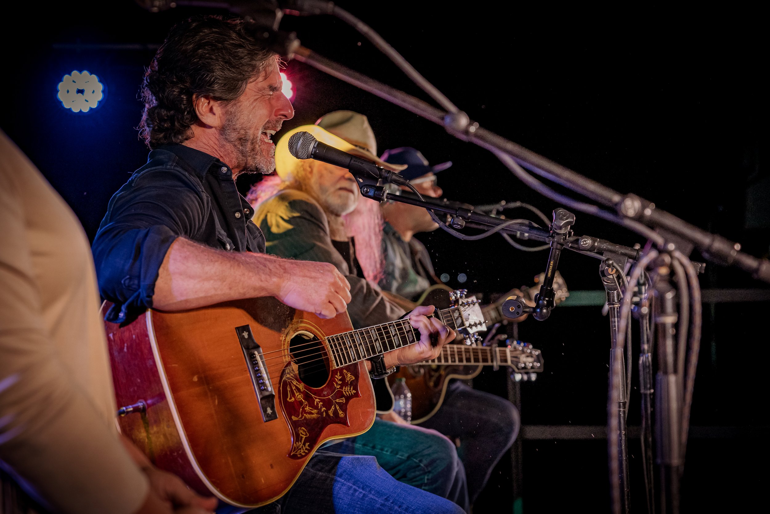 Four men playing acoustic guitars and singing into microphones on stage during a live performance.