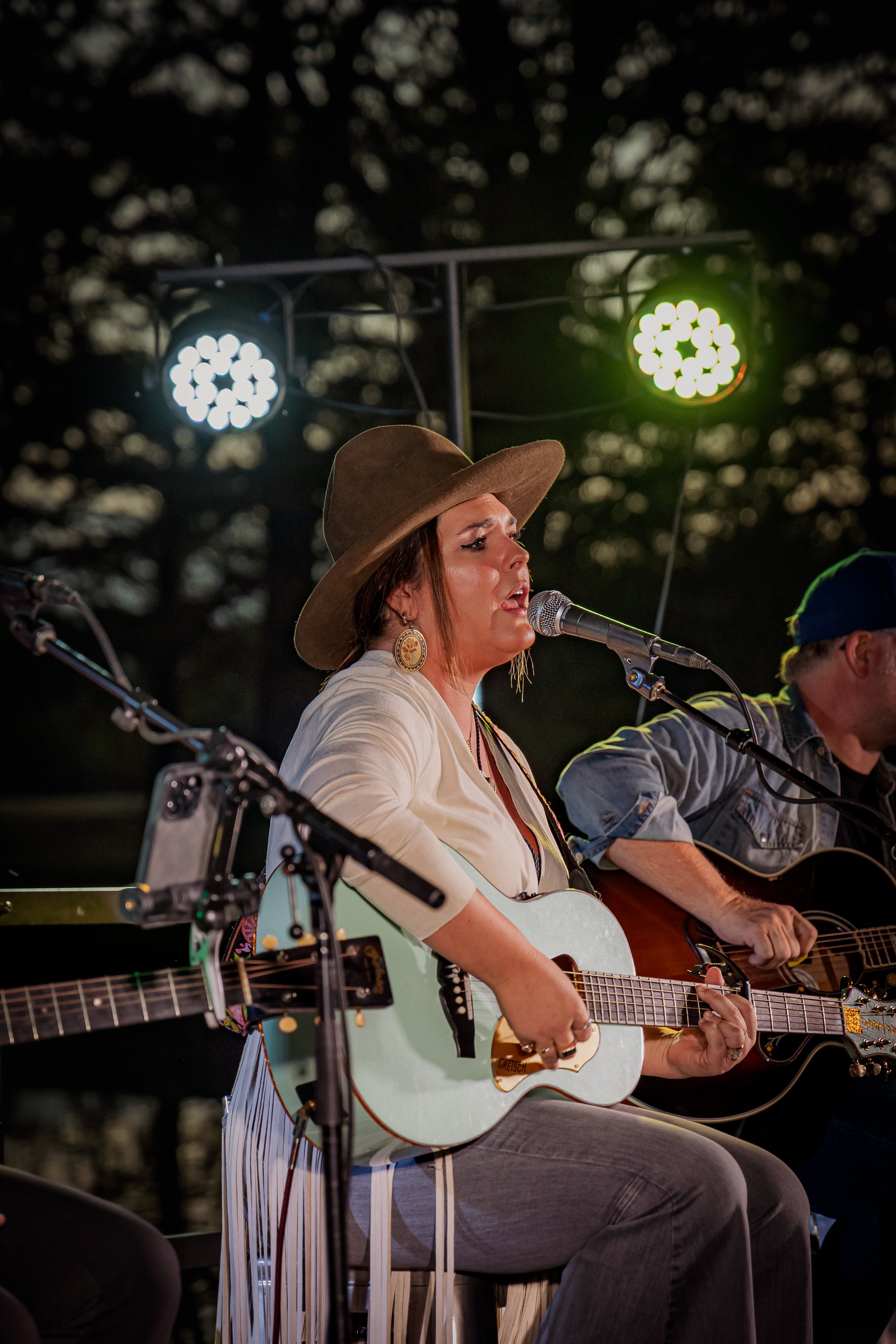 A woman wearing a wide-brimmed hat and earrings is singing into a microphone while playing an acoustic guitar. She is performing outdoors at dusk with stage lights overhead.